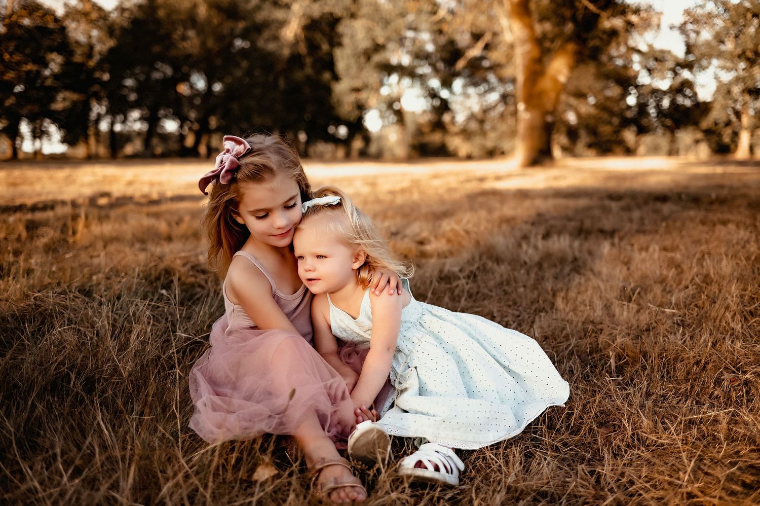 Two young girls in pretty dresses sitting together in a golden field at sunset, sharing a sweet sisterly moment.