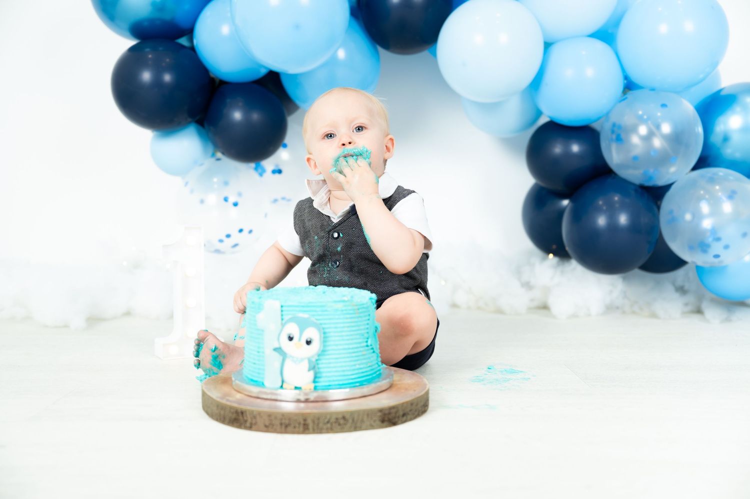 Baby boy smashing blue owl cake at first birthday party with navy and blue balloon arch backdrop.