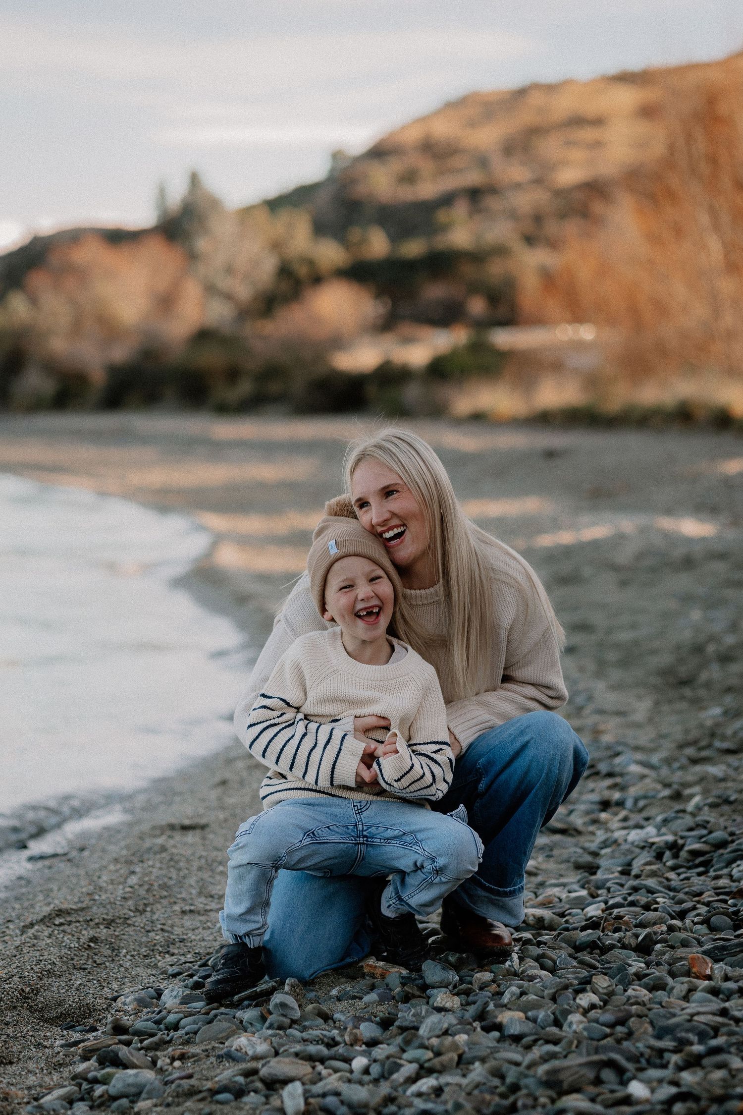 A smiling mother hugs her laughing child on a rocky beach with autumn cliffs in the background.