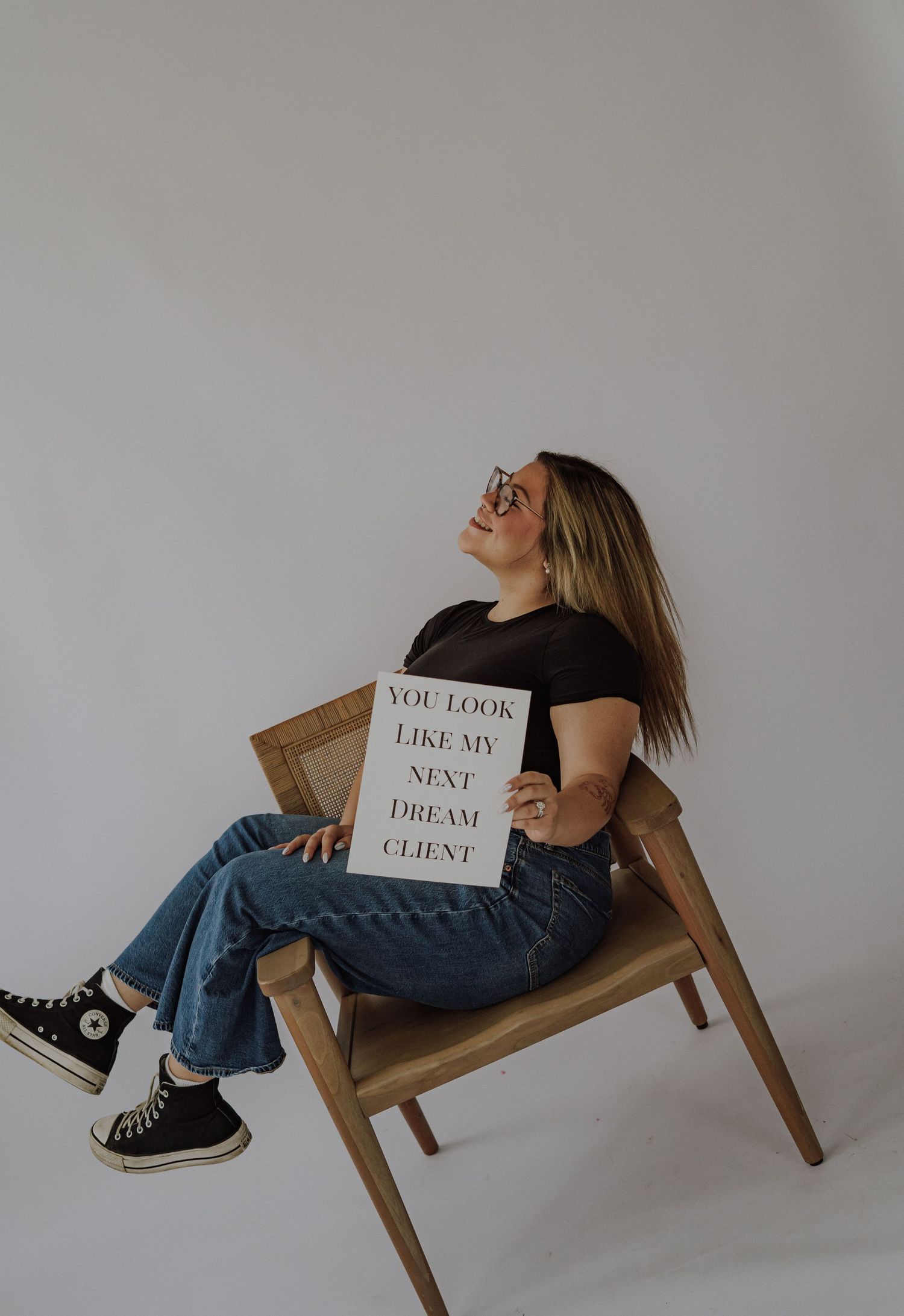 Photographer holding camera and 'Now Booking' sign in jeans and beige tee, promoting photography services.