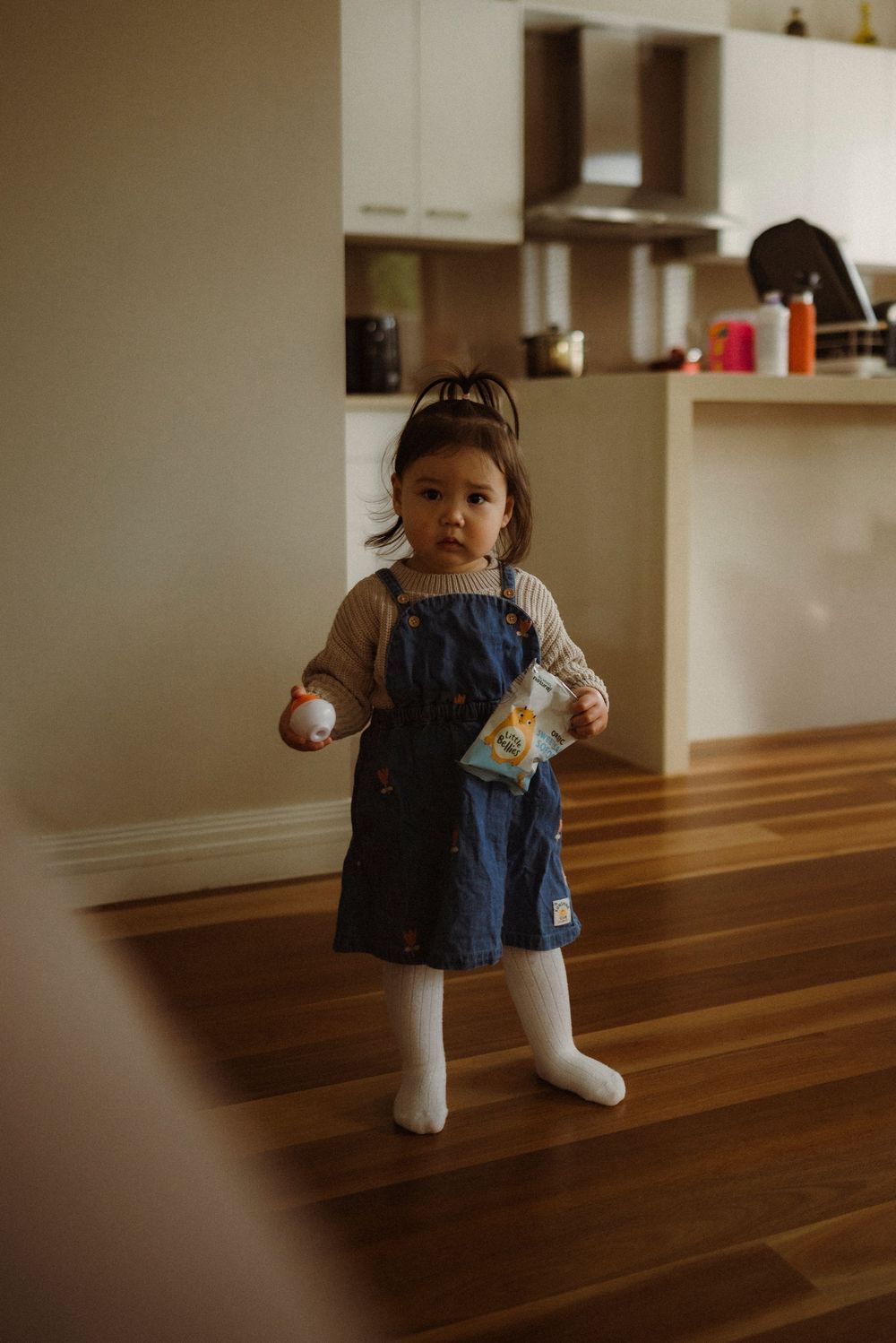 Cute toddler girl in denim overalls holding snacks standing on hardwood floor.