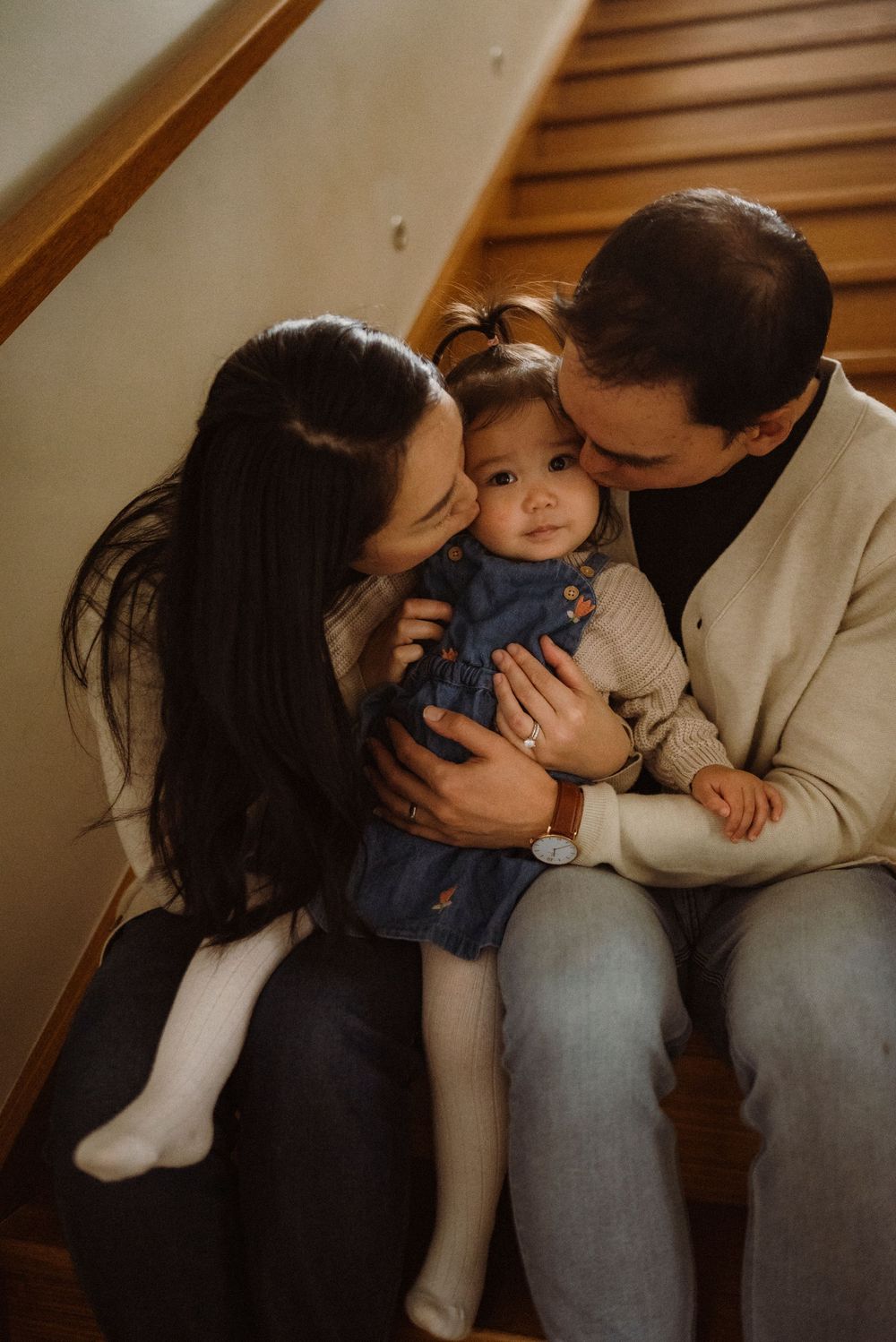 Loving parents sitting on wooden stairs kissing their baby girl on the cheeks.