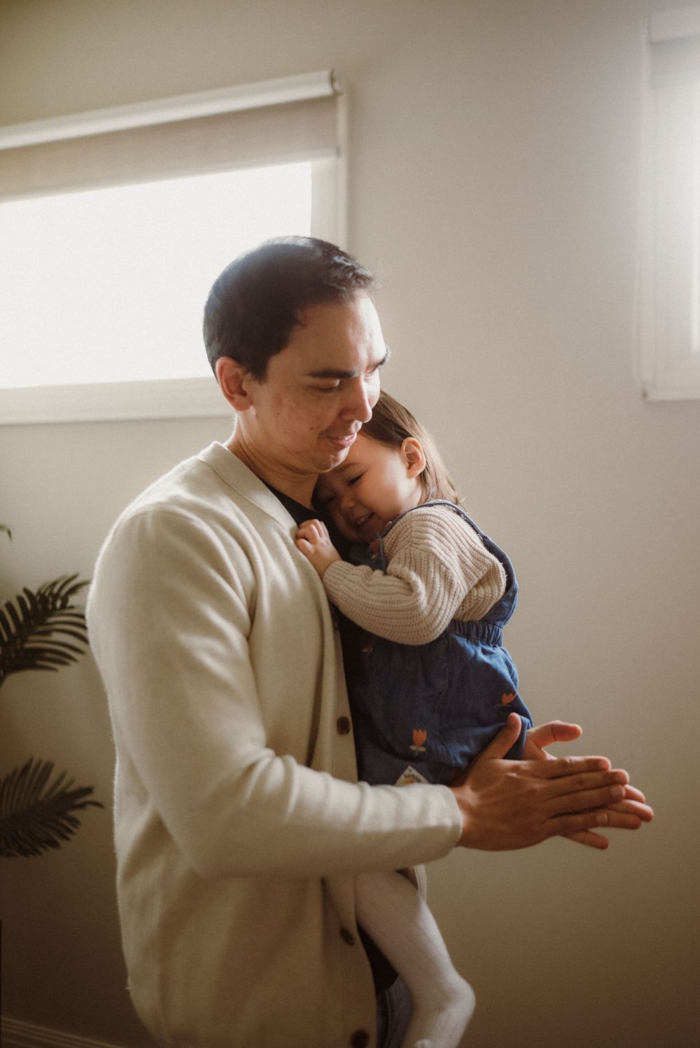 A father tenderly holds his young child close as soft natural light streams through a window behind them.