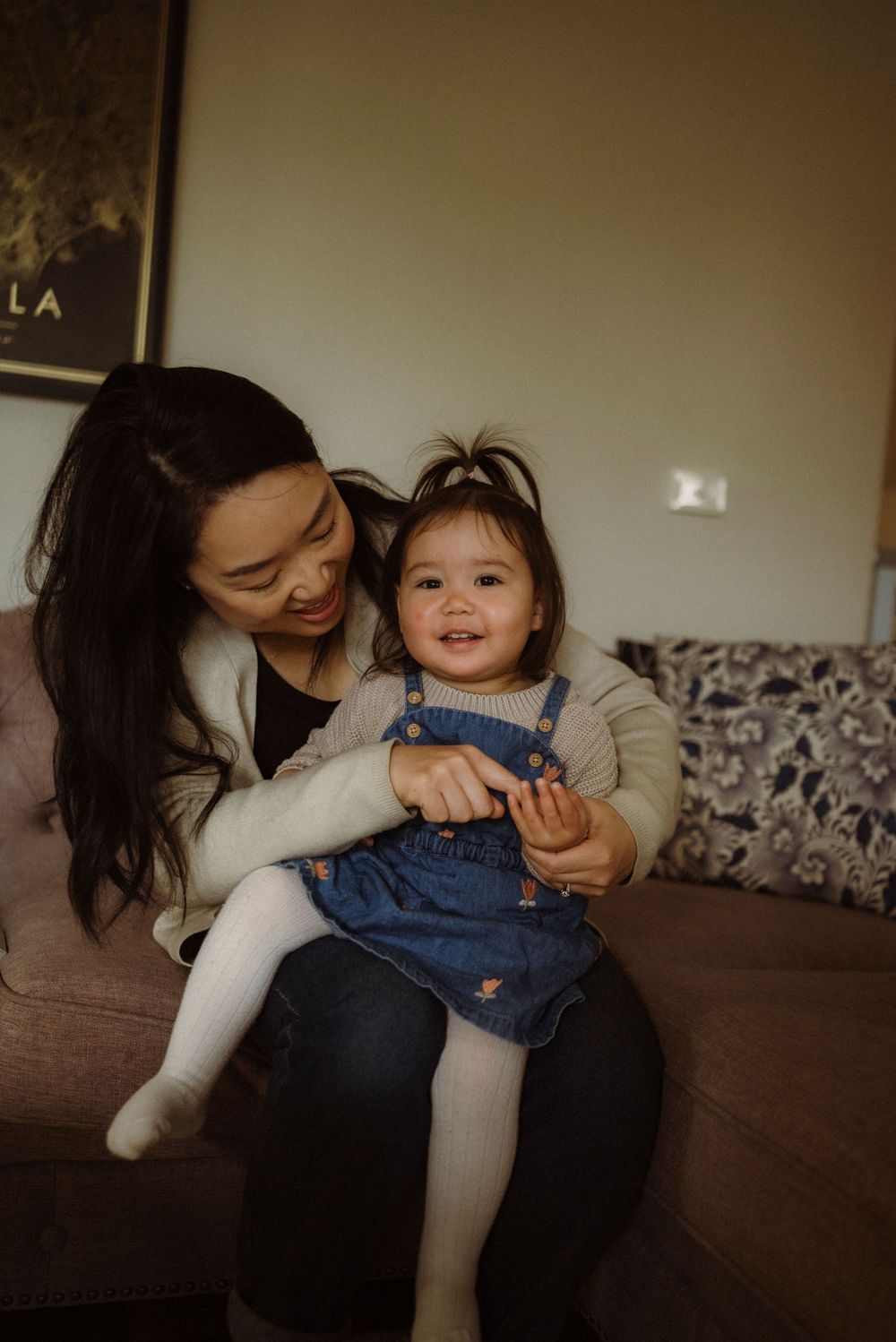 Mother holding her smiling toddler daughter on her lap while sitting on a couch.