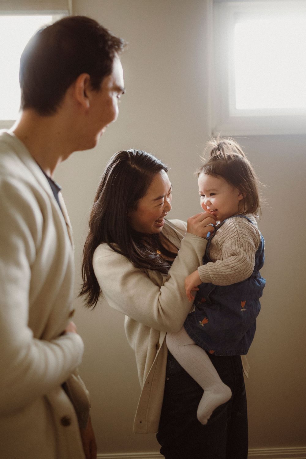 Asian mother holding toddler while father watches in a warmly lit room.