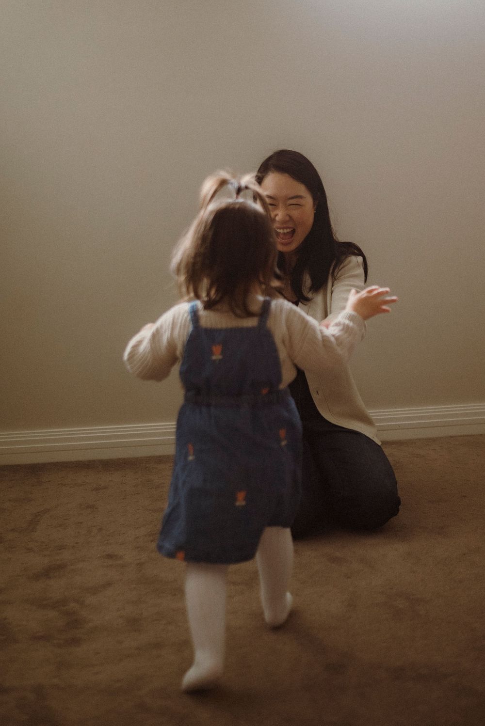 A mother kneels on carpet, arms open wide, laughing as her toddler girl in blue denim dress runs toward her.