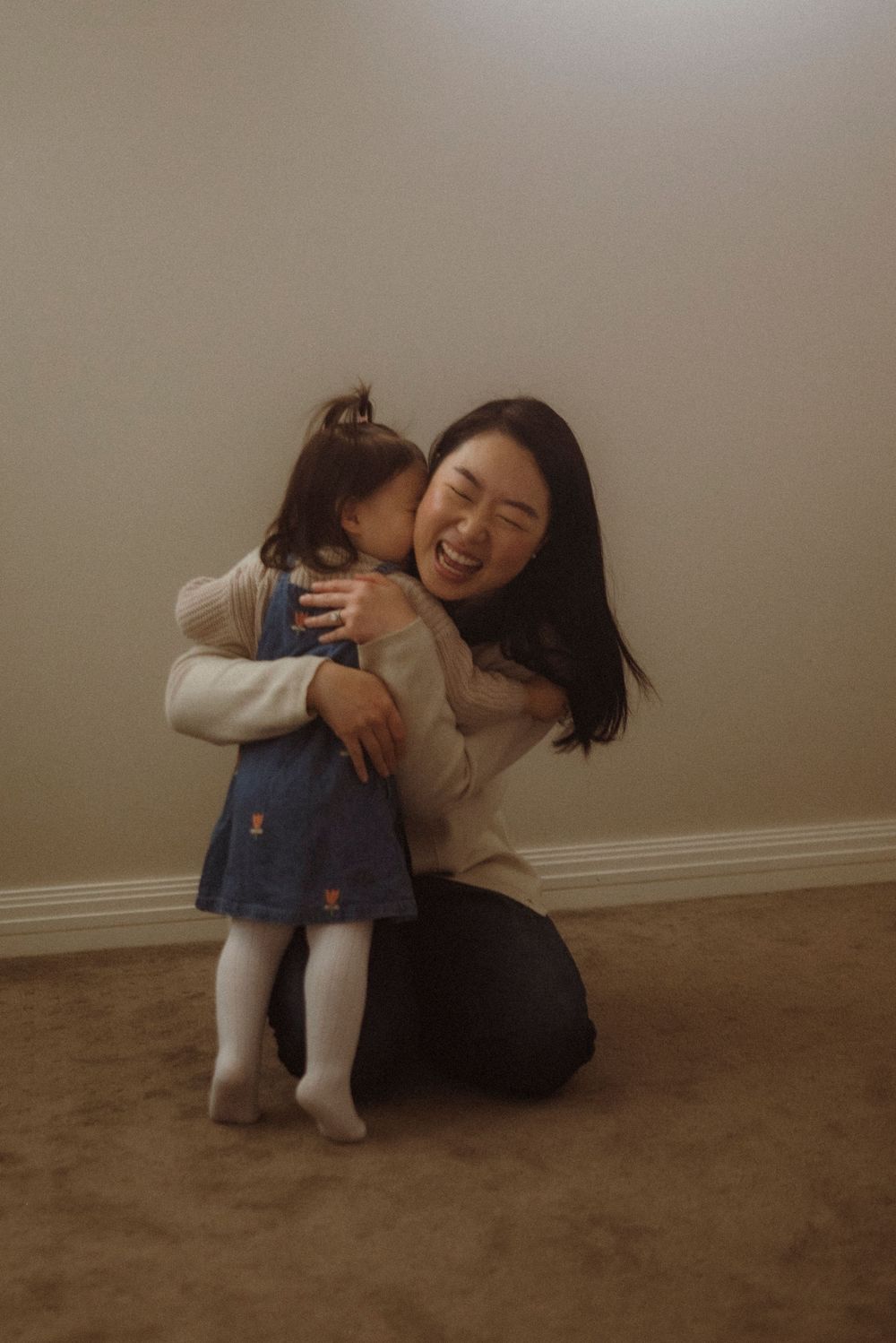 A laughing Asian woman kneels on the floor hugging a toddler girl in a denim dress and white tights.