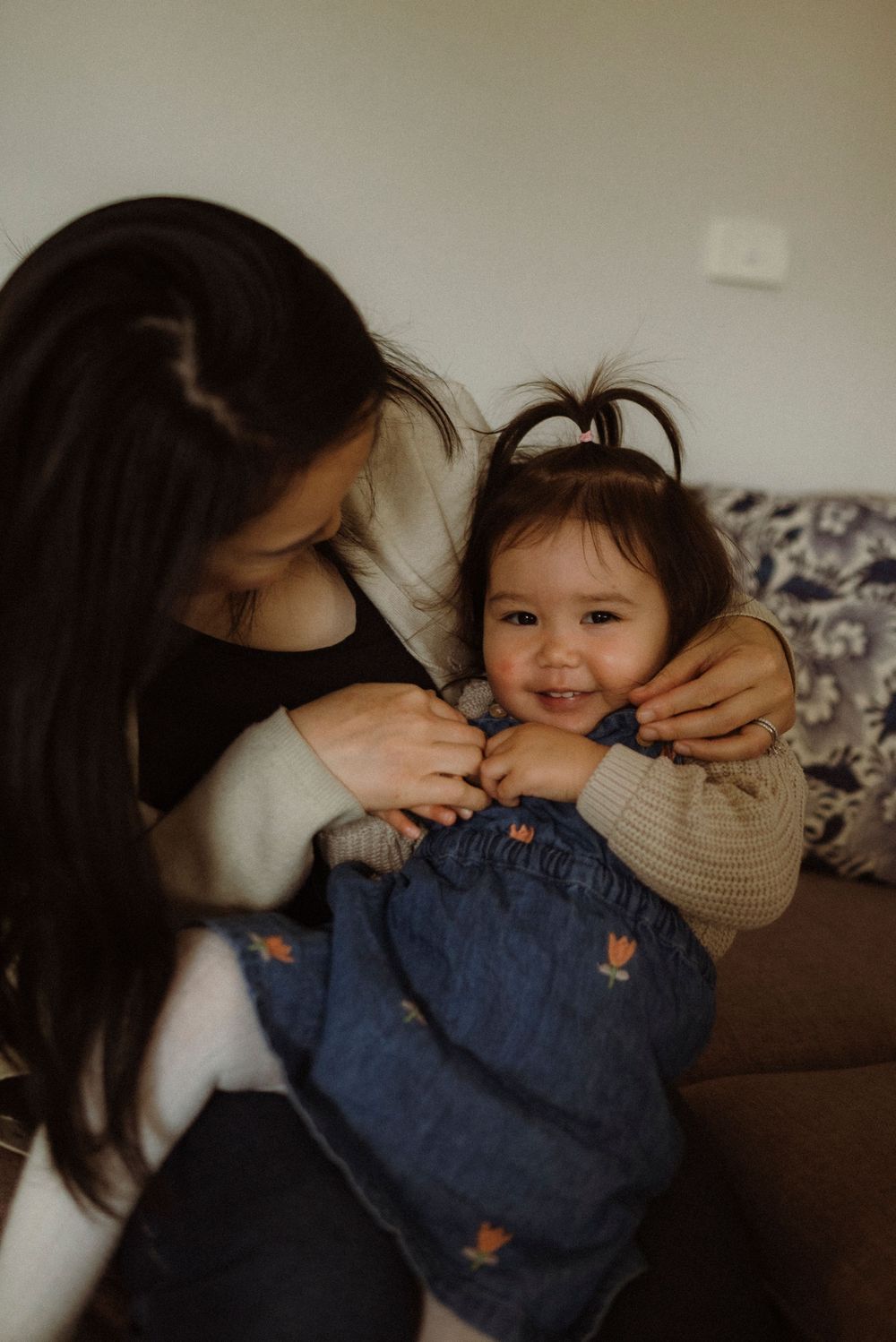 A mother lovingly holds her smiling toddler girl on a couch, both wearing casual clothes in a warm indoor setting.
