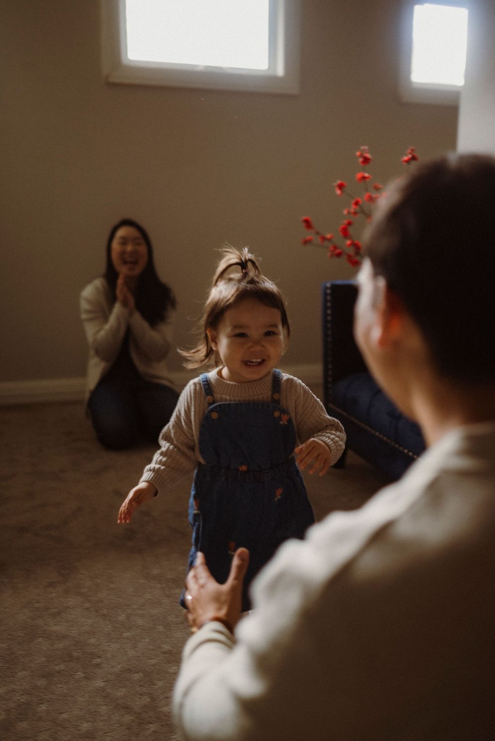 Happy toddler girl in denim overalls runs toward her father while mother laughs in the background of a cozy room.
