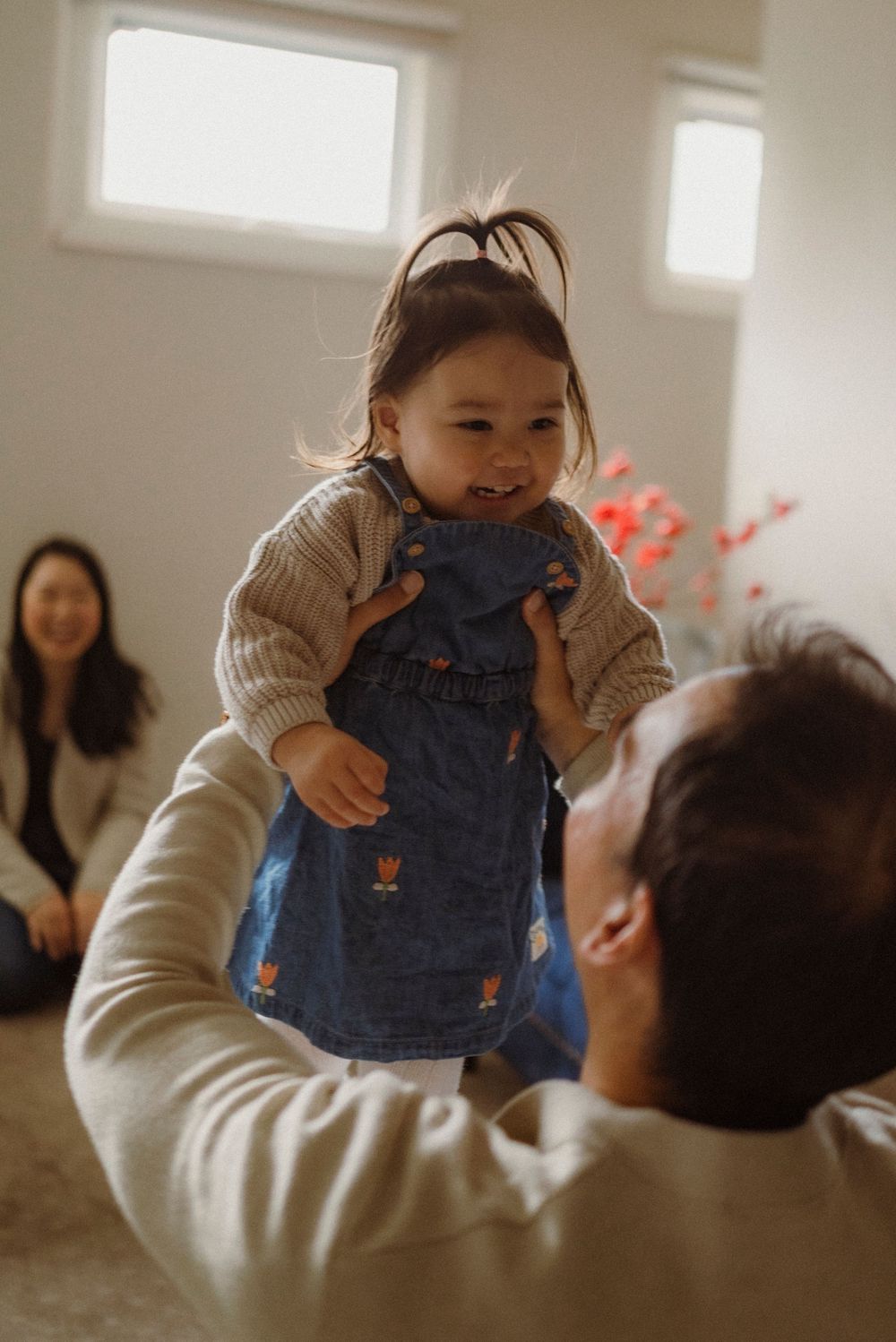 A father lifts his smiling toddler girl in a denim dress while a woman laughs in the background.