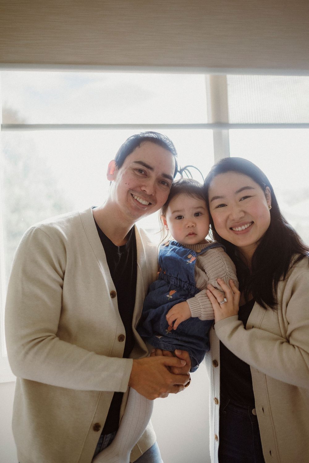 Smiling family portrait with parents holding their baby girl, dressed in coordinating cream cardigans.