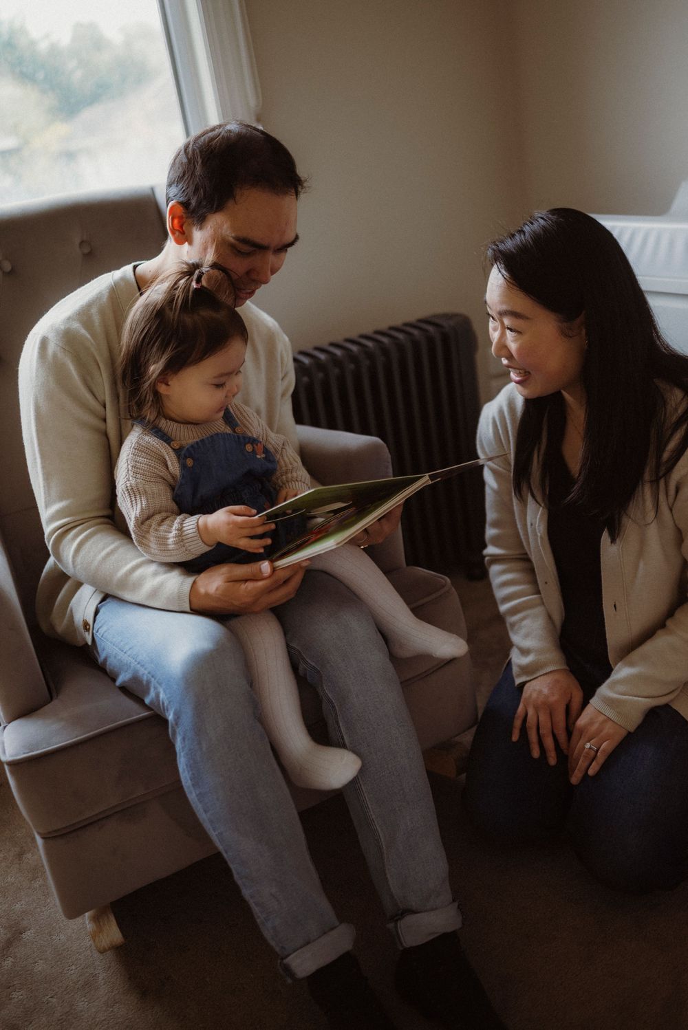 A family reads a picture book together, with a toddler on dad's lap and mom smiling beside them in a cozy room.