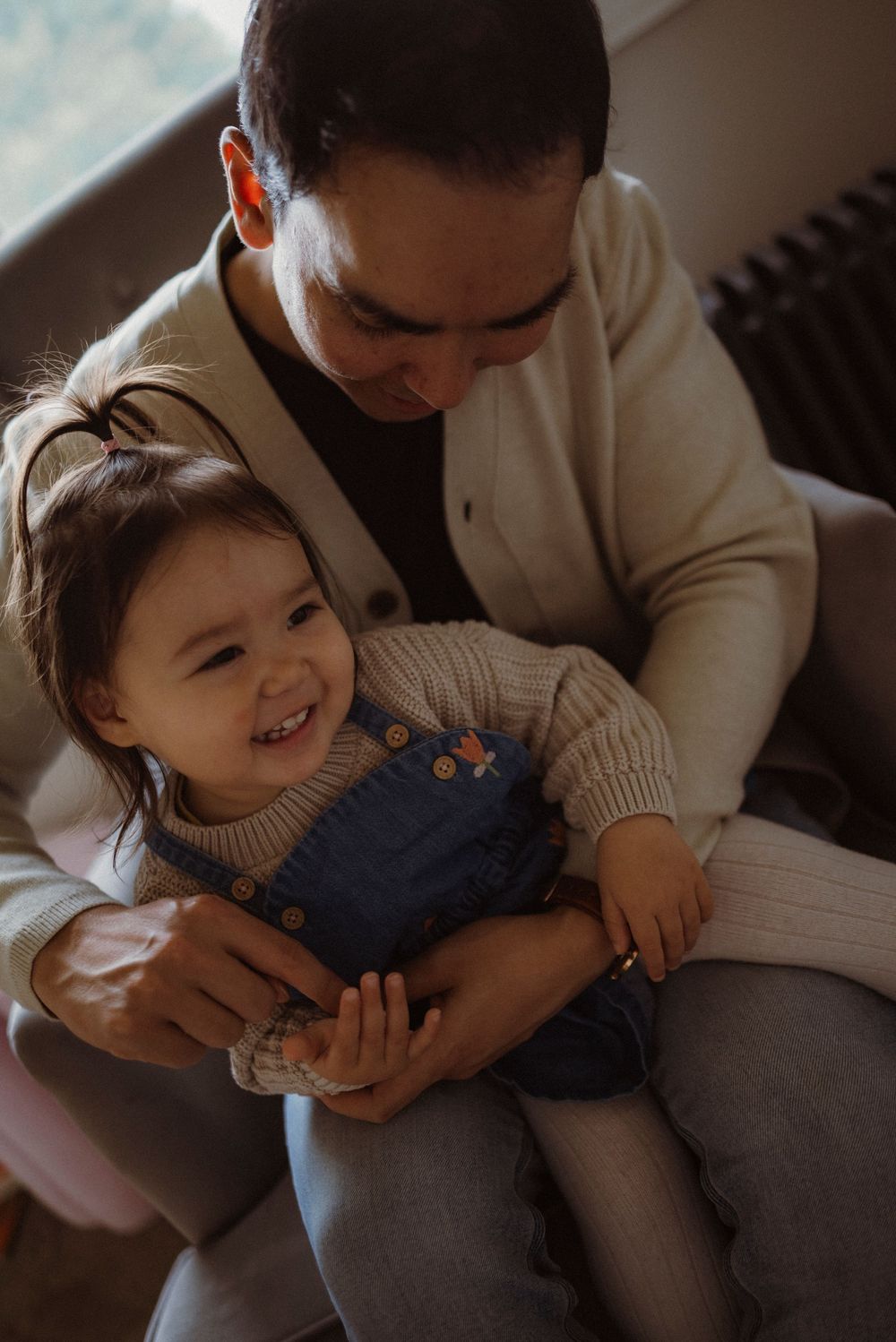 A smiling toddler in denim overalls sits on an adult's lap, sharing a warm moment indoors.