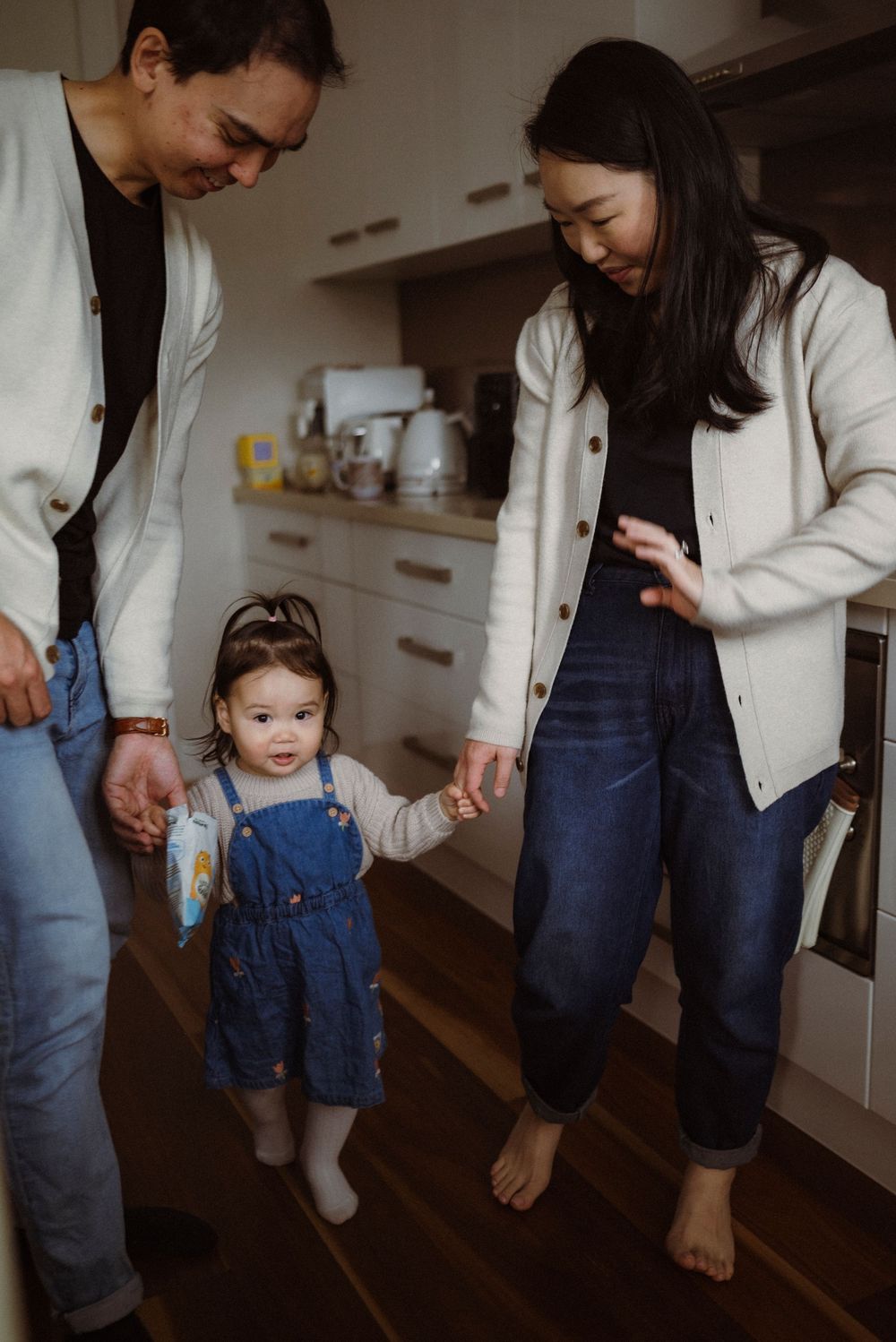 Two parents hold hands with their toddler daughter in a blue denim dress while standing in a modern kitchen.