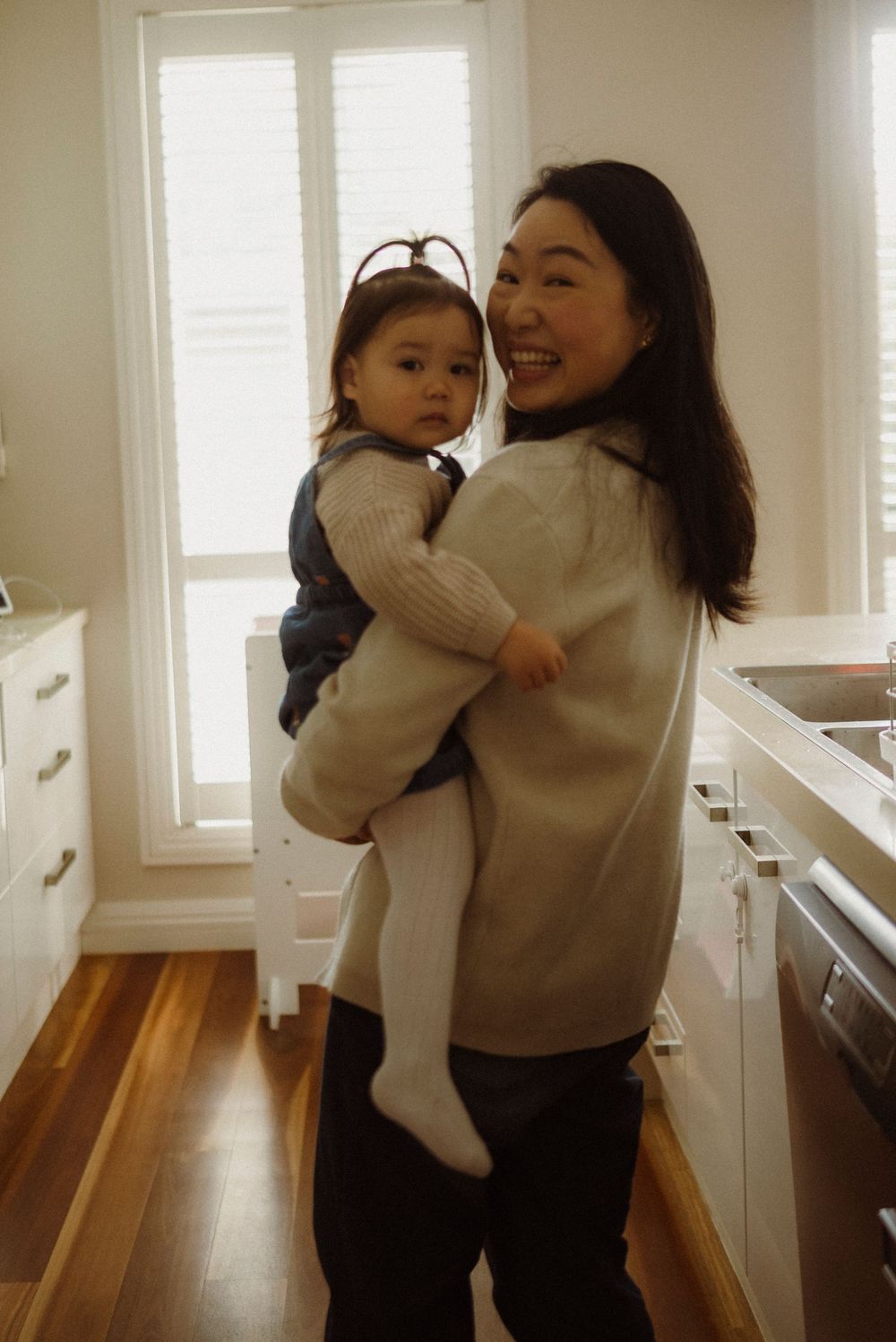 Smiling Asian mother holding her baby girl in a bright white kitchen with hardwood floors.