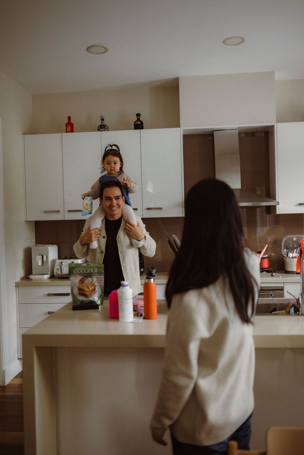 Father with toddler on shoulders in white kitchen while mother watches from island.