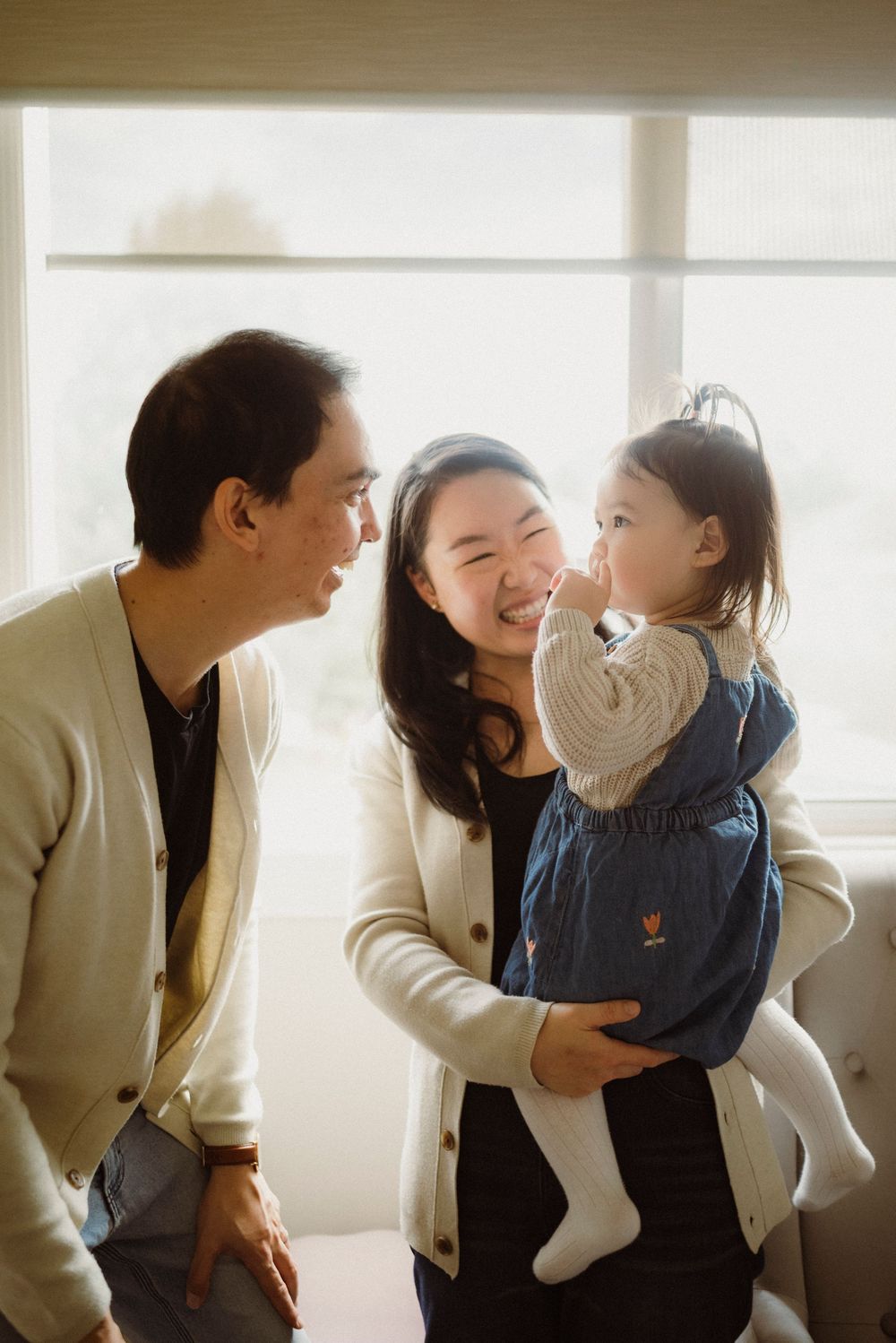 Happy Asian family with mother holding smiling toddler girl in denim dress while father looks on lovingly.