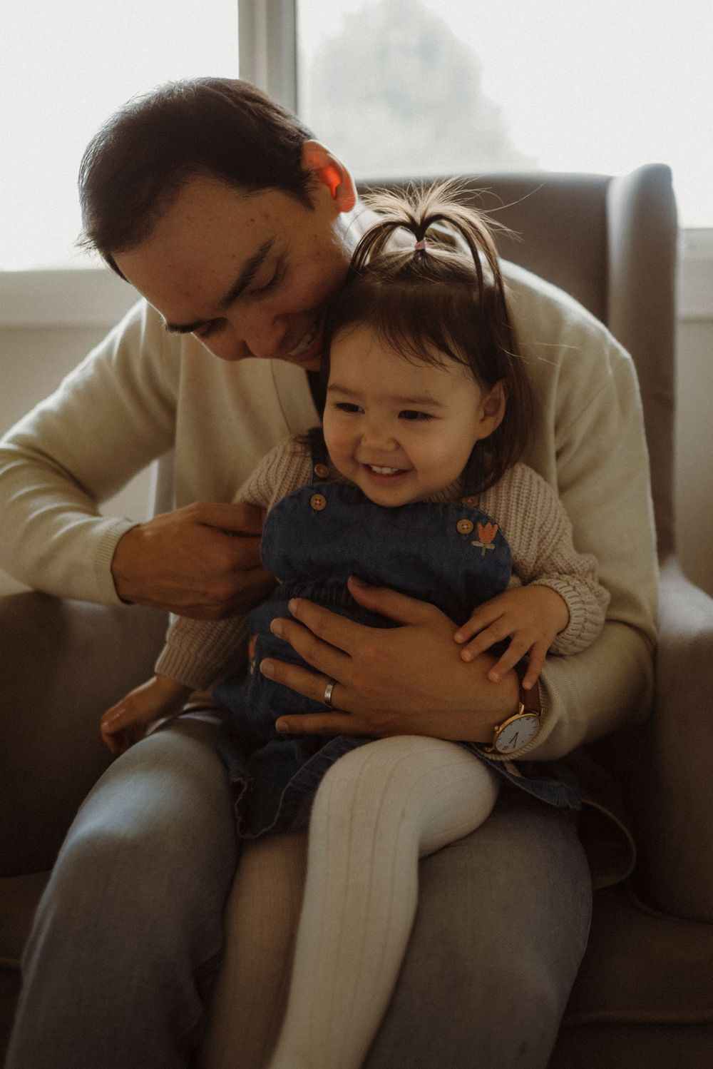 A father cuddles his giggling toddler daughter in a denim dress on a cozy armchair by a bright window.