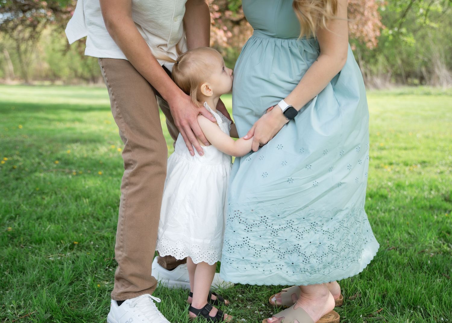 Pregnant woman in mint dress with toddler and partner standing on green grass in outdoor maternity photo.