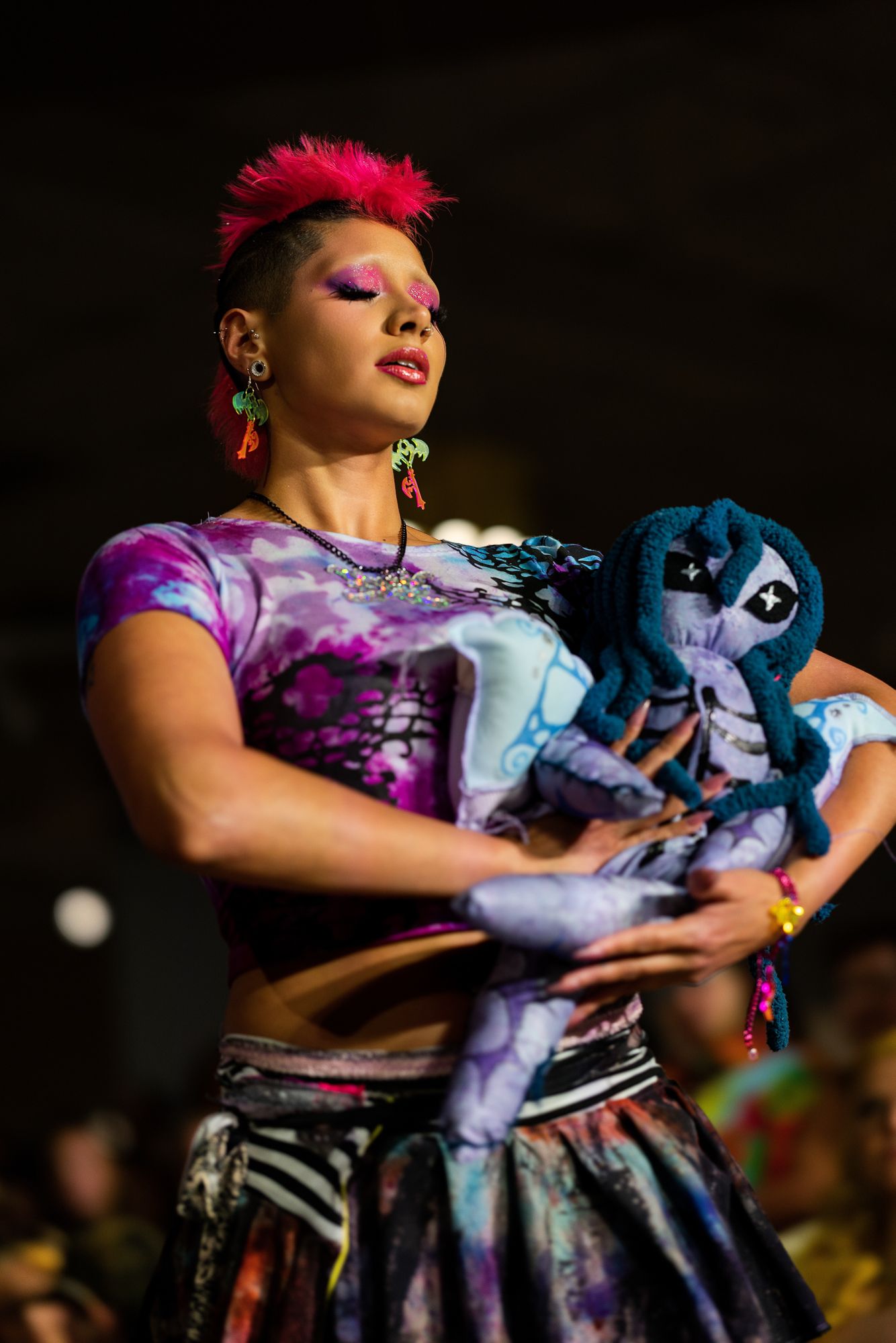 Colorful performer with pink hair holds a handmade doll on a fashion runway stage.