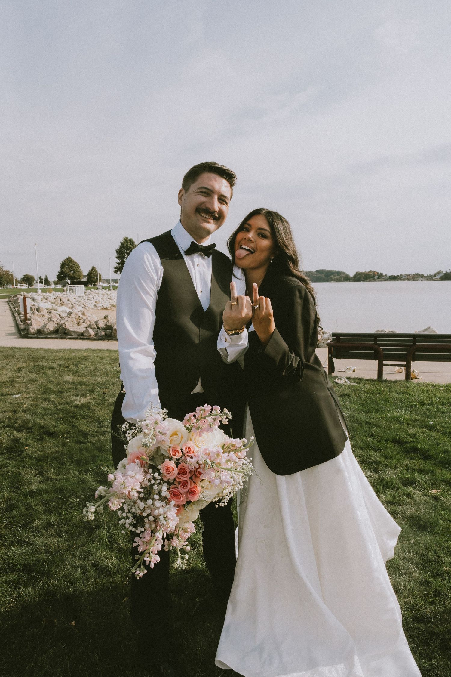 Smiling newlyweds pose outdoors near waterfront, bride in white gown showing wedding ring, groom holding pink floral bouquet.