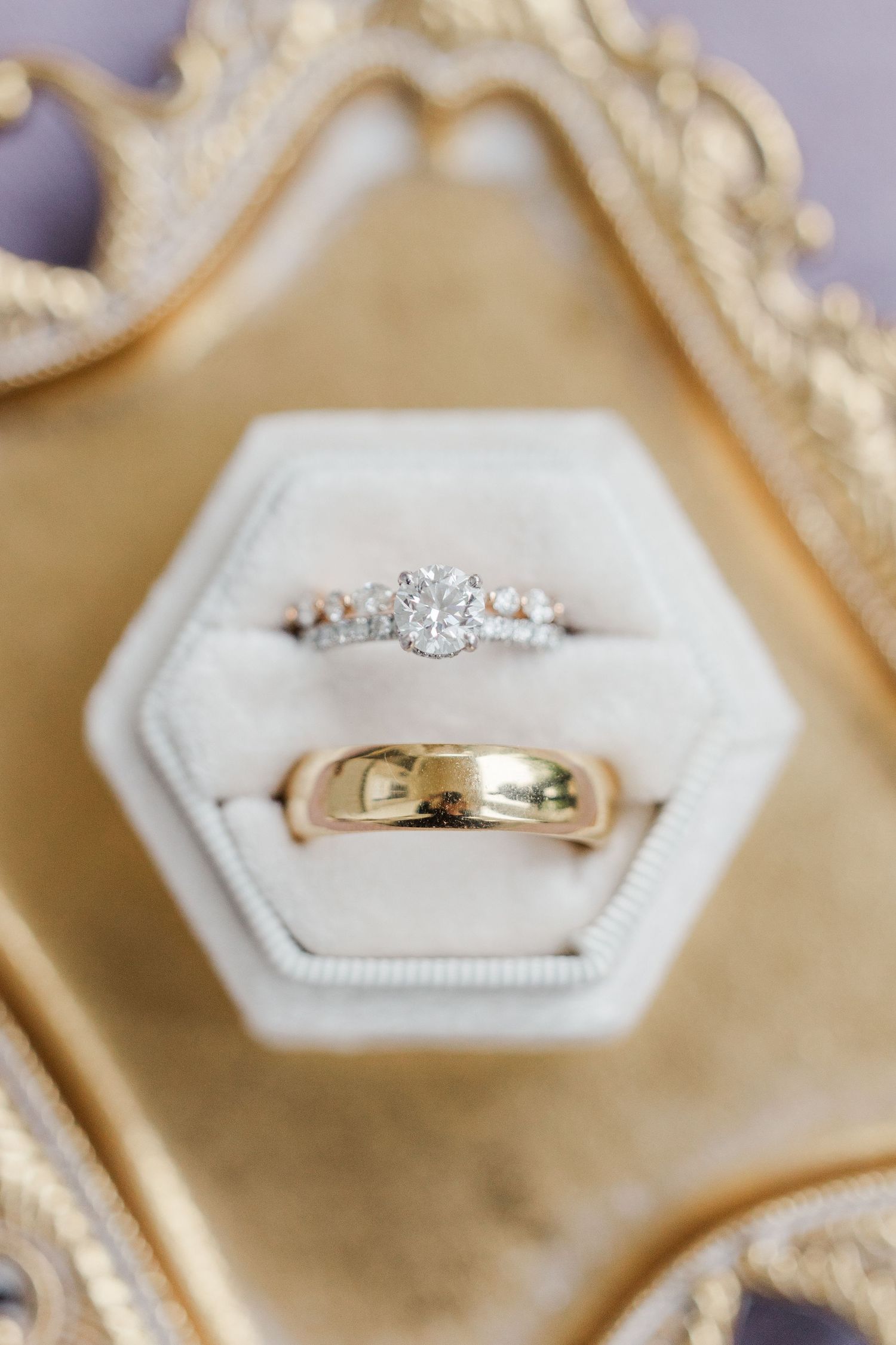 Diamond engagement ring and gold wedding band displayed in a white velvet hexagon ring box on a gold tray.