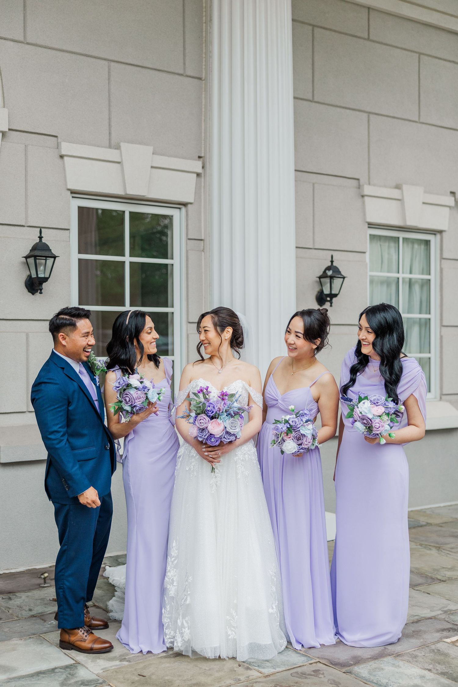 Bride in white lace gown with bridesmaids in lavender dresses holding purple bouquets outside elegant venue.