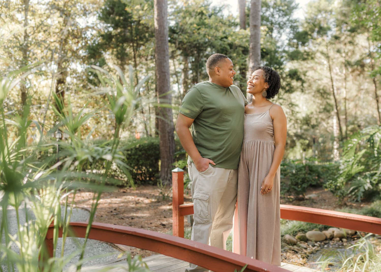 Couple standing on a wooden bridge surrounded by lush green trees, sharing a romantic moment outdoors.