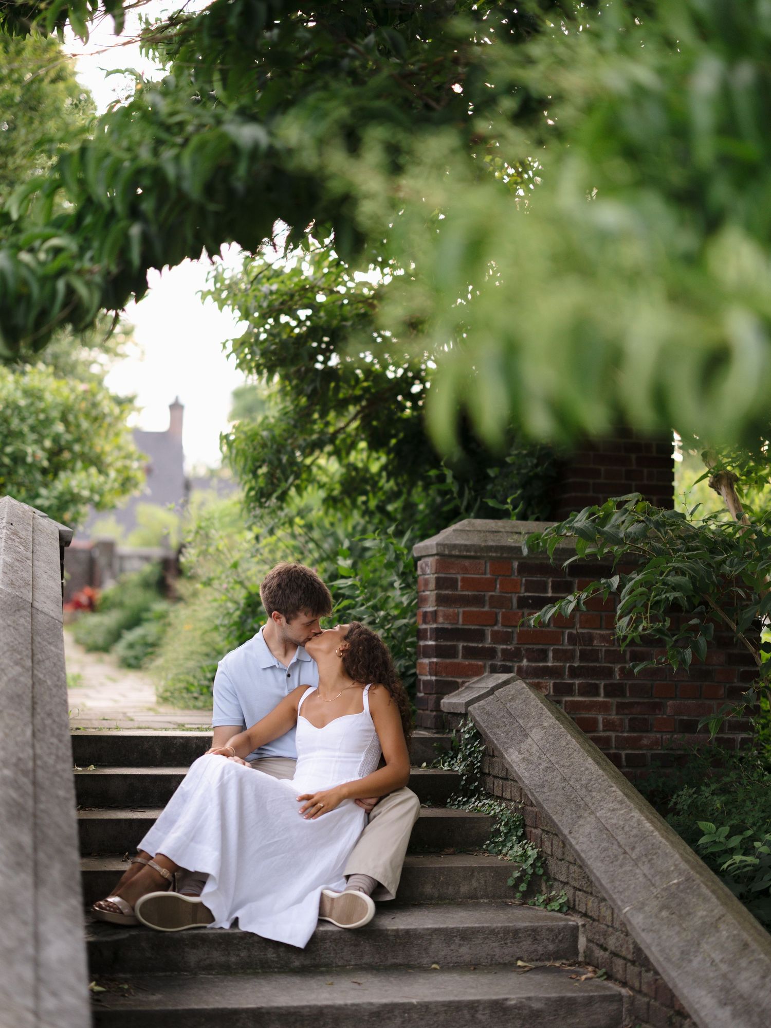 Couple sharing a tender kiss while sitting on stone steps surrounded by green trees and brick walls.