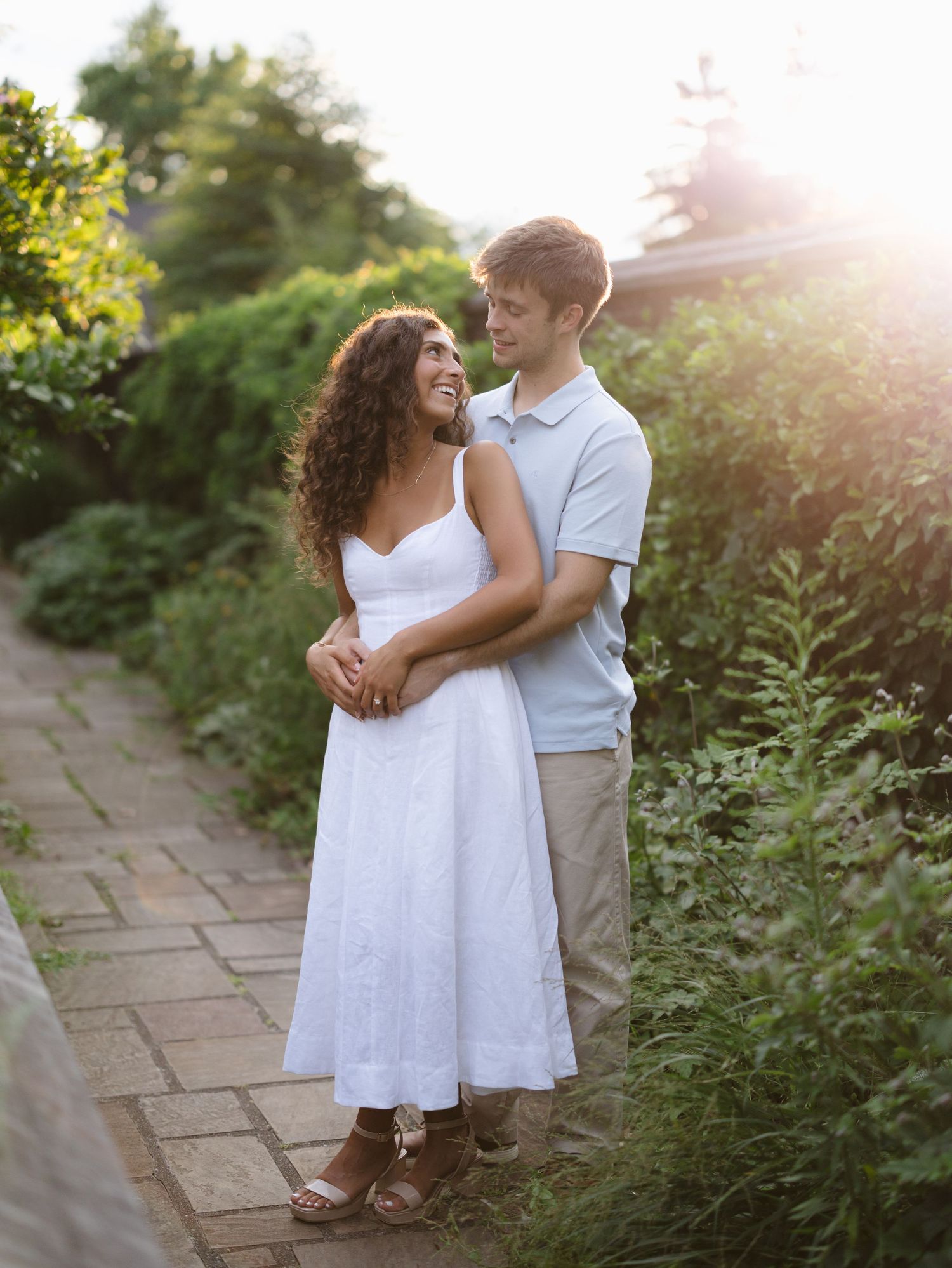 A couple embraces on a brick garden path, smiling at each other in golden sunset light surrounded by lush greenery.