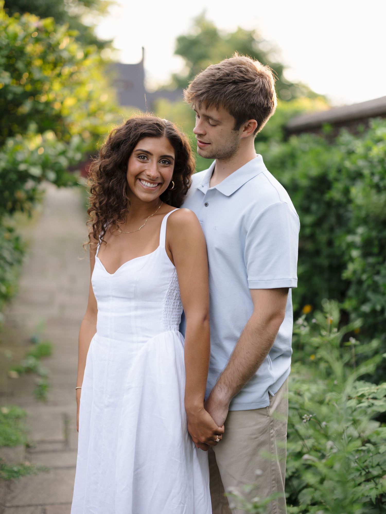 Smiling couple posing together on a garden path surrounded by greenery, woman in white dress.