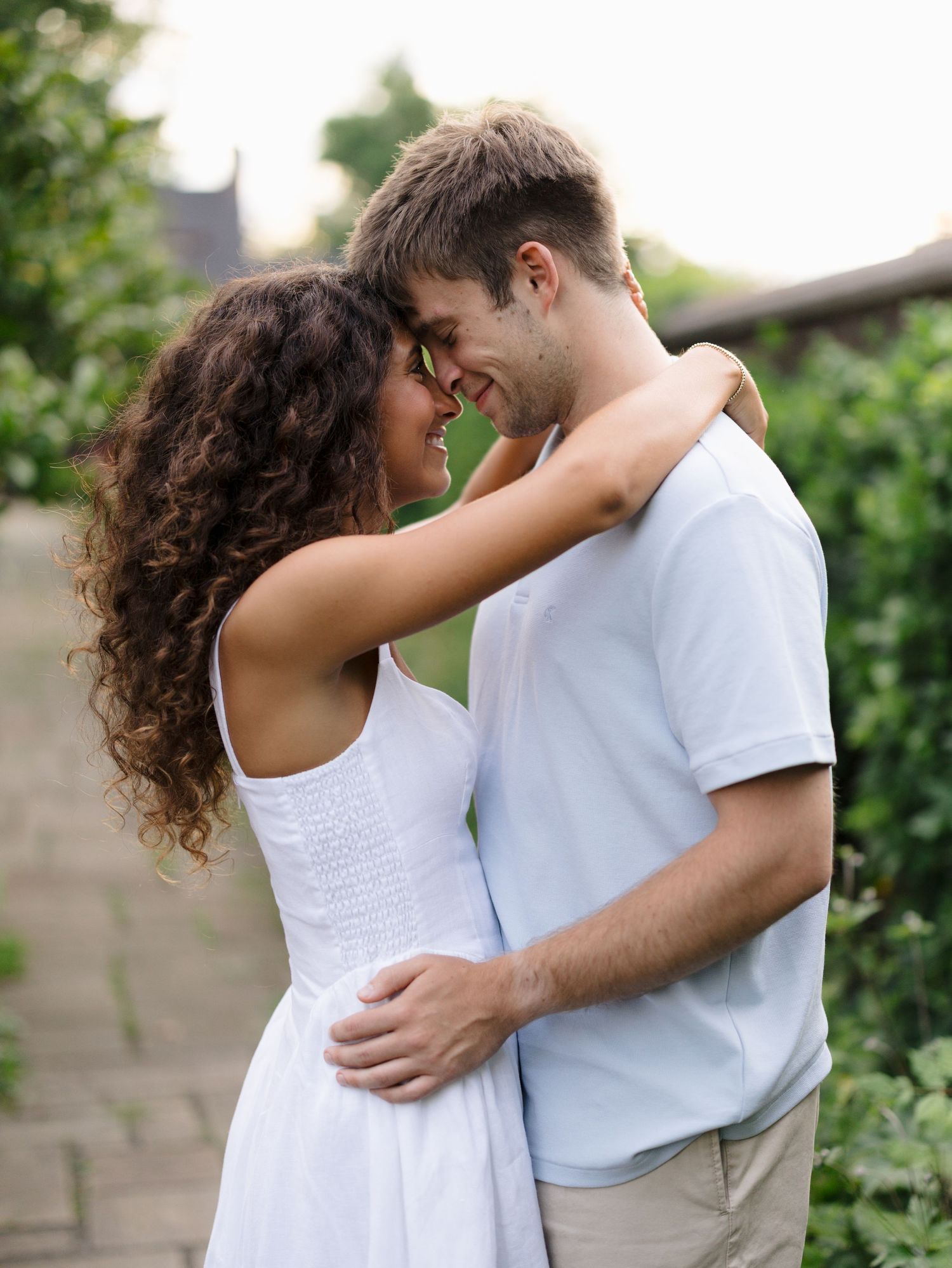 A romantic couple embracing outdoors, smiling at each other in a lush green garden setting, wearing casual white outfits.