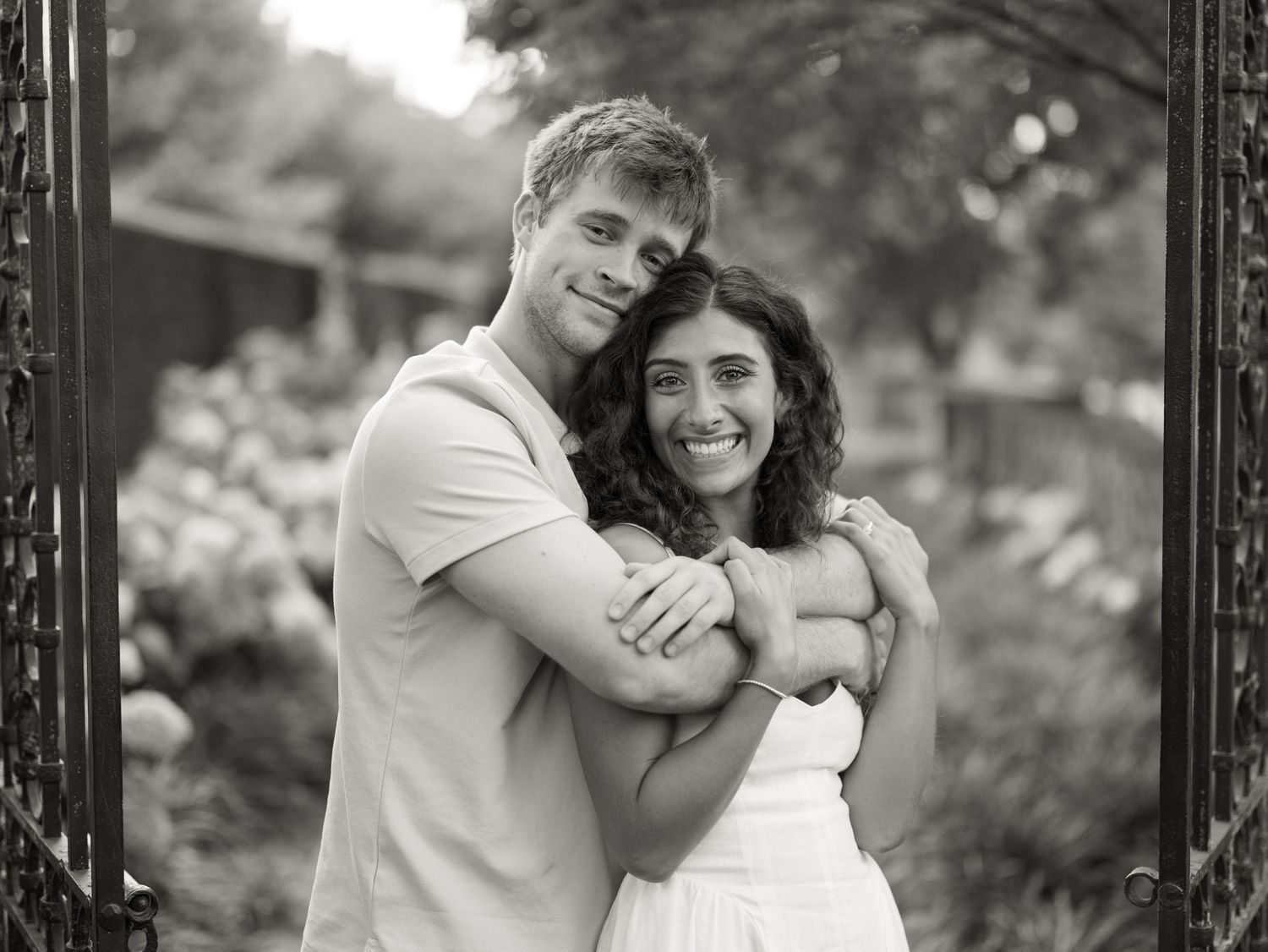 Black and white photo of a smiling couple embracing outdoors, man hugging woman in white dress near garden gate.