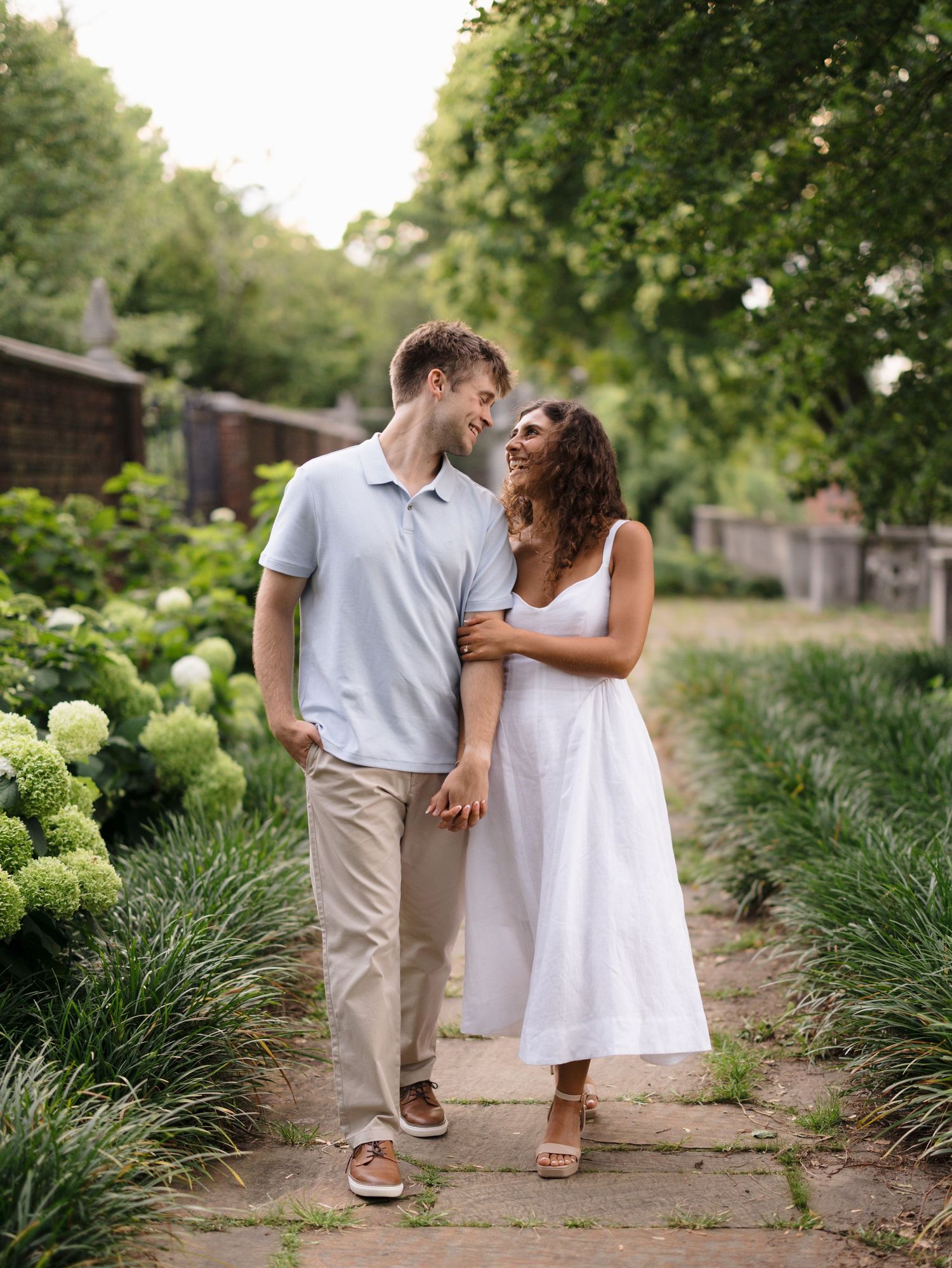 A smiling couple walks hand-in-hand along a garden path surrounded by lush greenery and white hydrangeas.