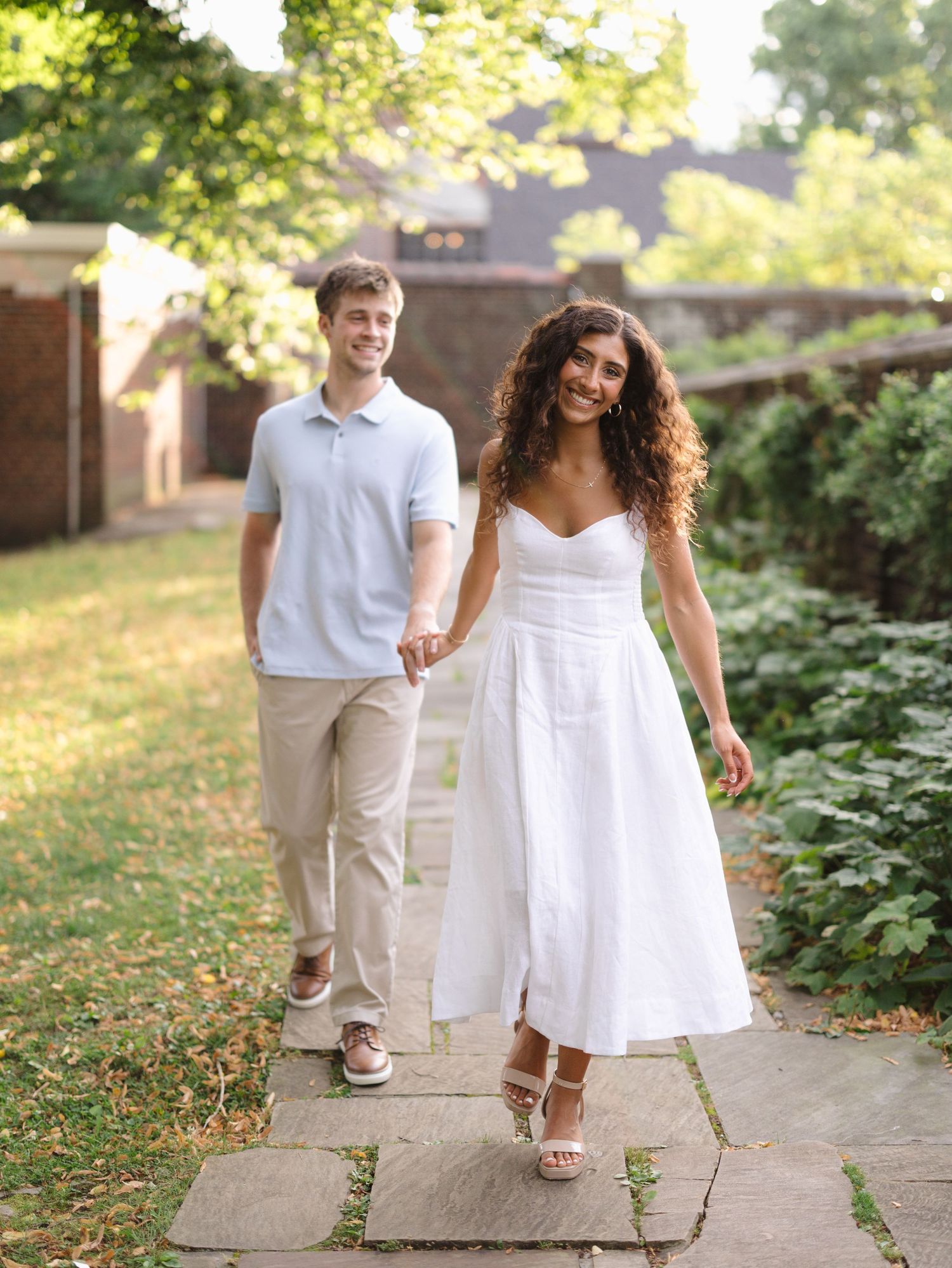 A couple walks hand-in-hand on a garden path, woman in white dress and man in light blue polo.