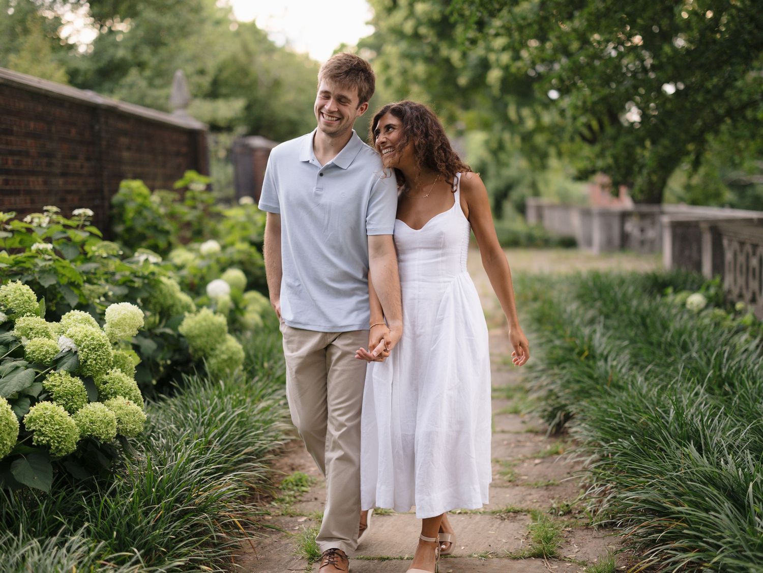Smiling couple strolling hand in hand along garden path lined with hydrangeas, dressed in casual summer attire.