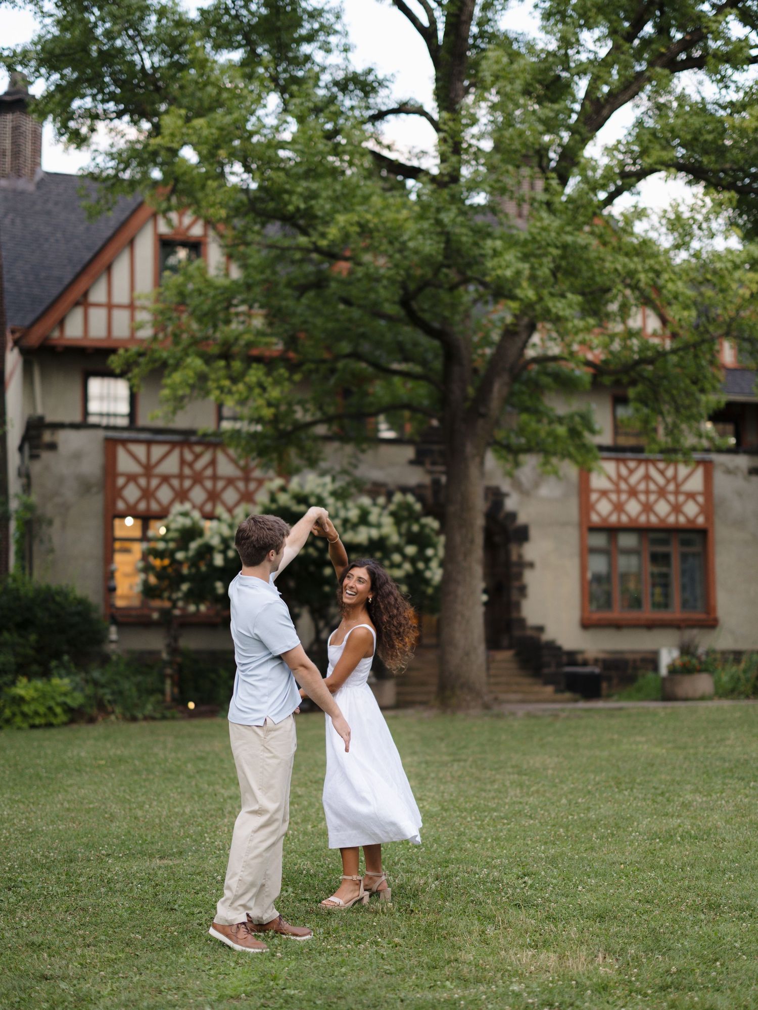 Couple dancing on green lawn in front of Tudor-style mansion with large oak tree at sunset.