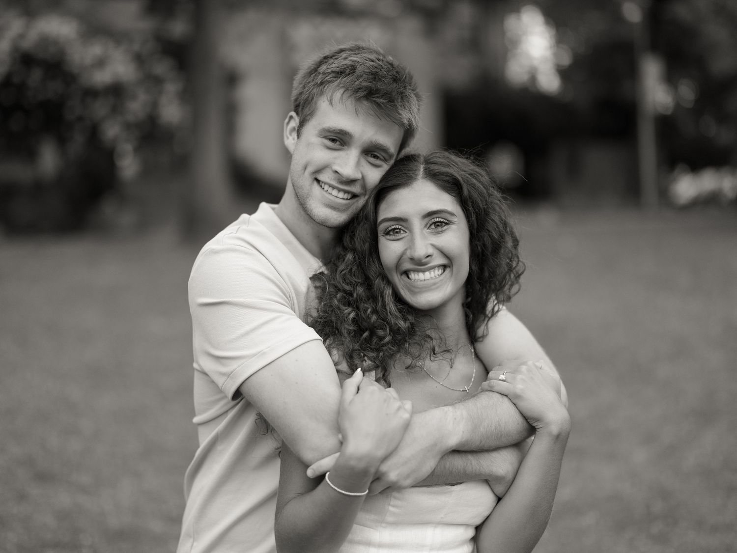 Black and white photo of a smiling couple embracing outdoors, man hugging woman from behind in a park setting.