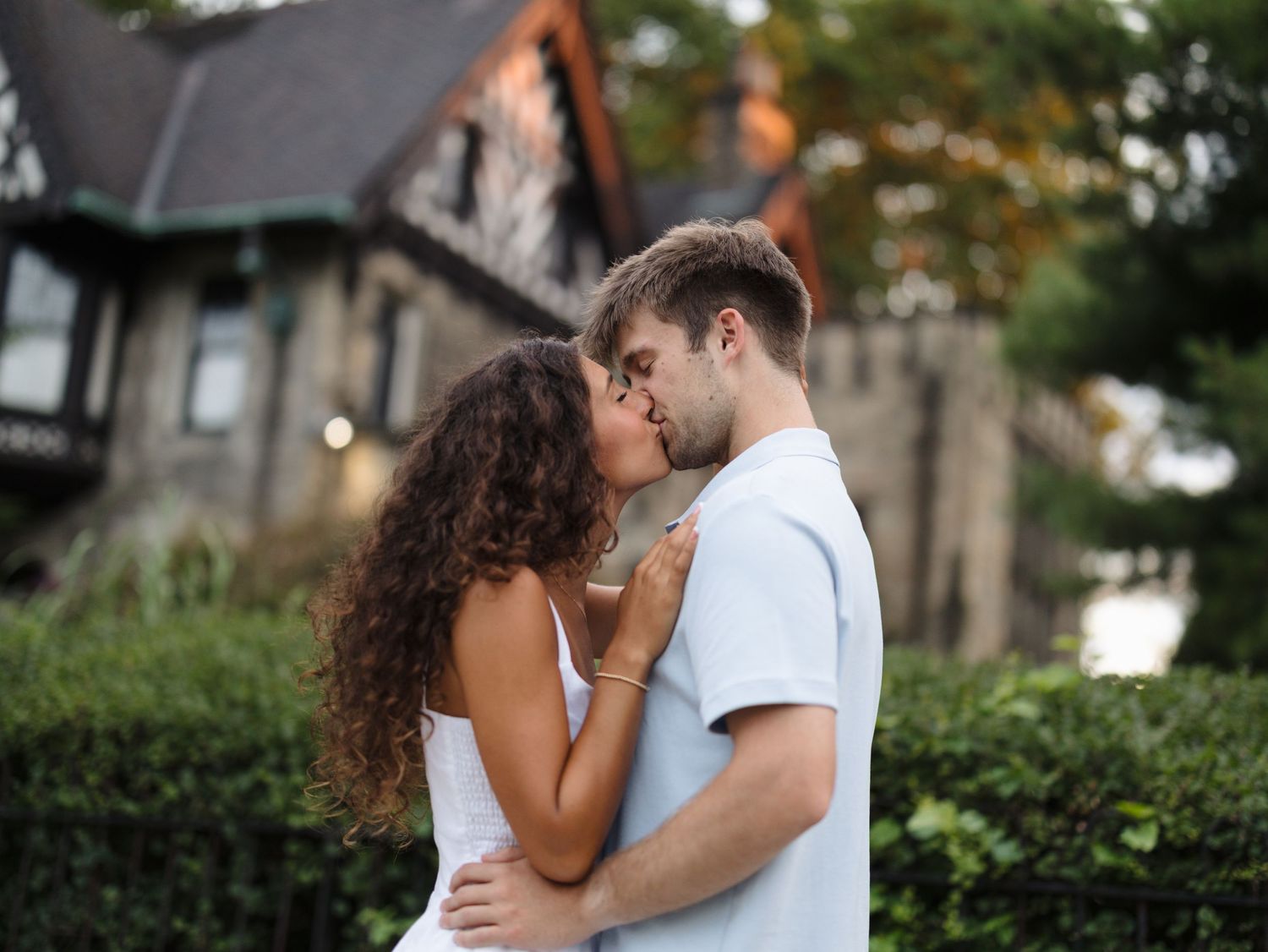 A couple shares a romantic kiss outdoors in front of a charming stone house surrounded by lush green hedges.