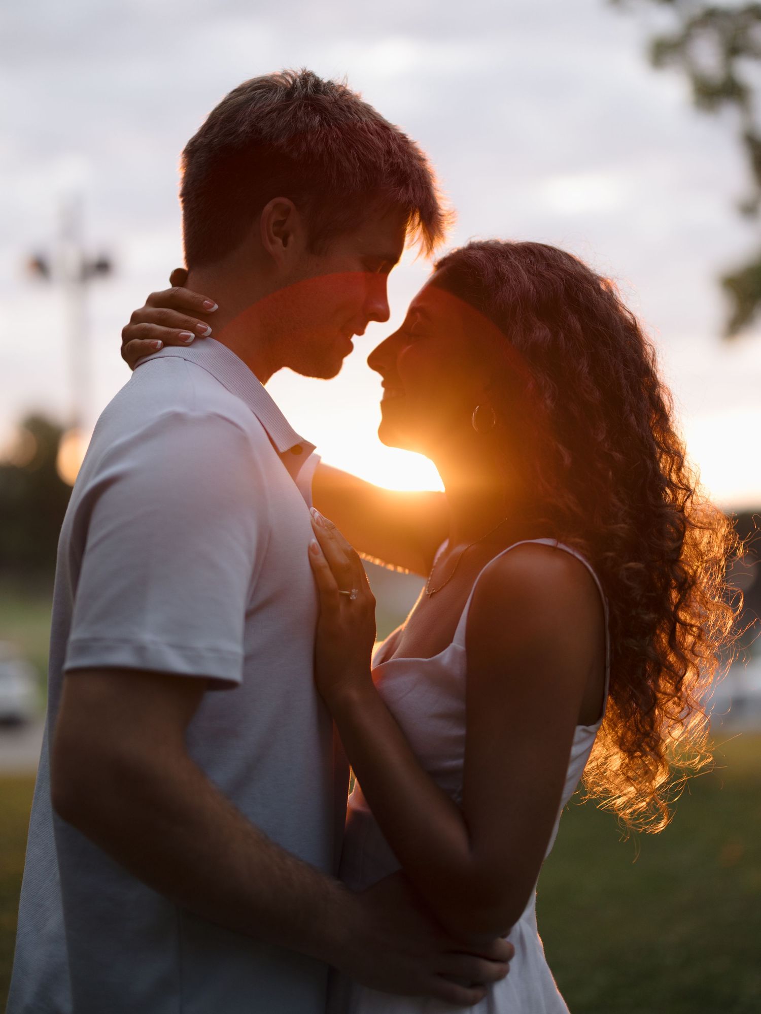 Romantic couple embracing at golden sunset in a park, backlit by warm sunlight with the woman's curly hair glowing.