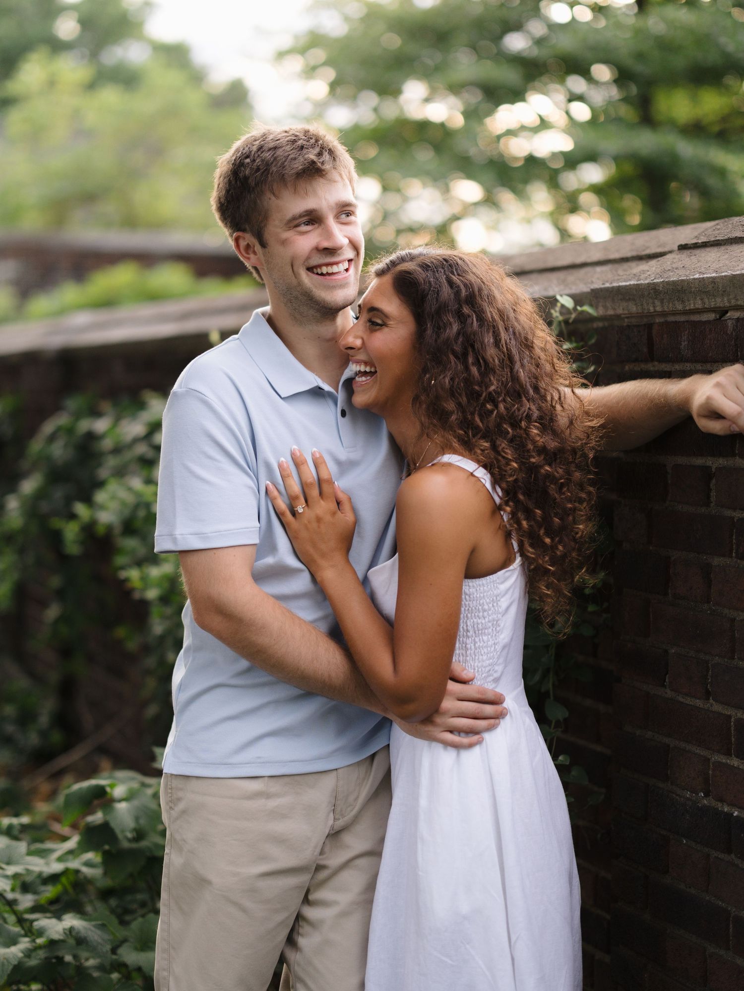 Happy couple embracing outdoors near brick wall, woman in white dress showing engagement ring, lush green trees in background.