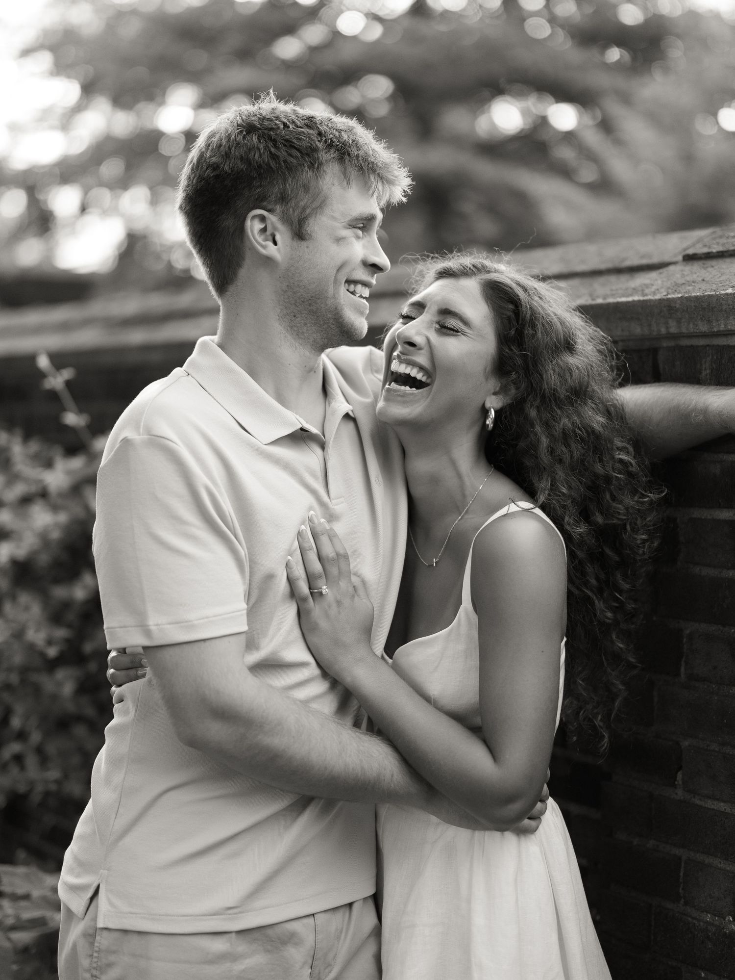 Black and white photo of a laughing couple embracing against a brick wall in a lush garden setting.