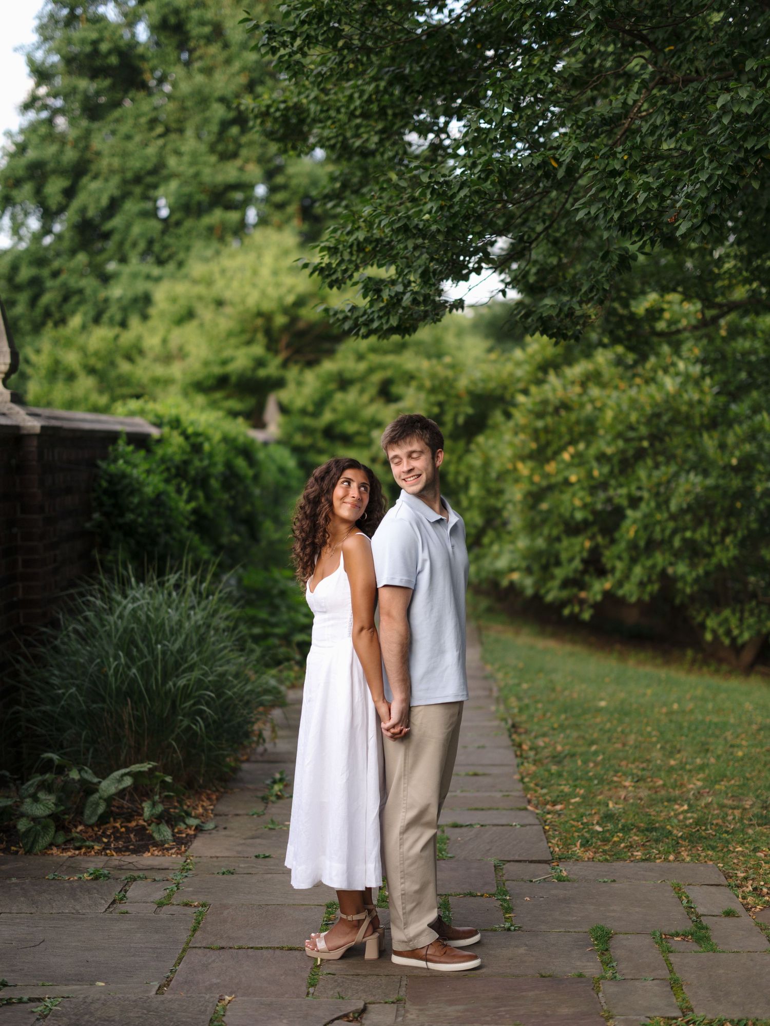 Couple holding hands on a stone garden path surrounded by lush green trees and shrubs.