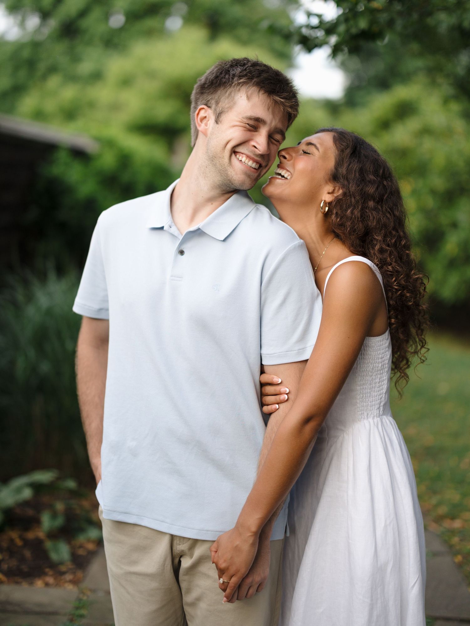 A smiling couple embraces outdoors, woman in white dress leaning close to laughing man in blue polo.