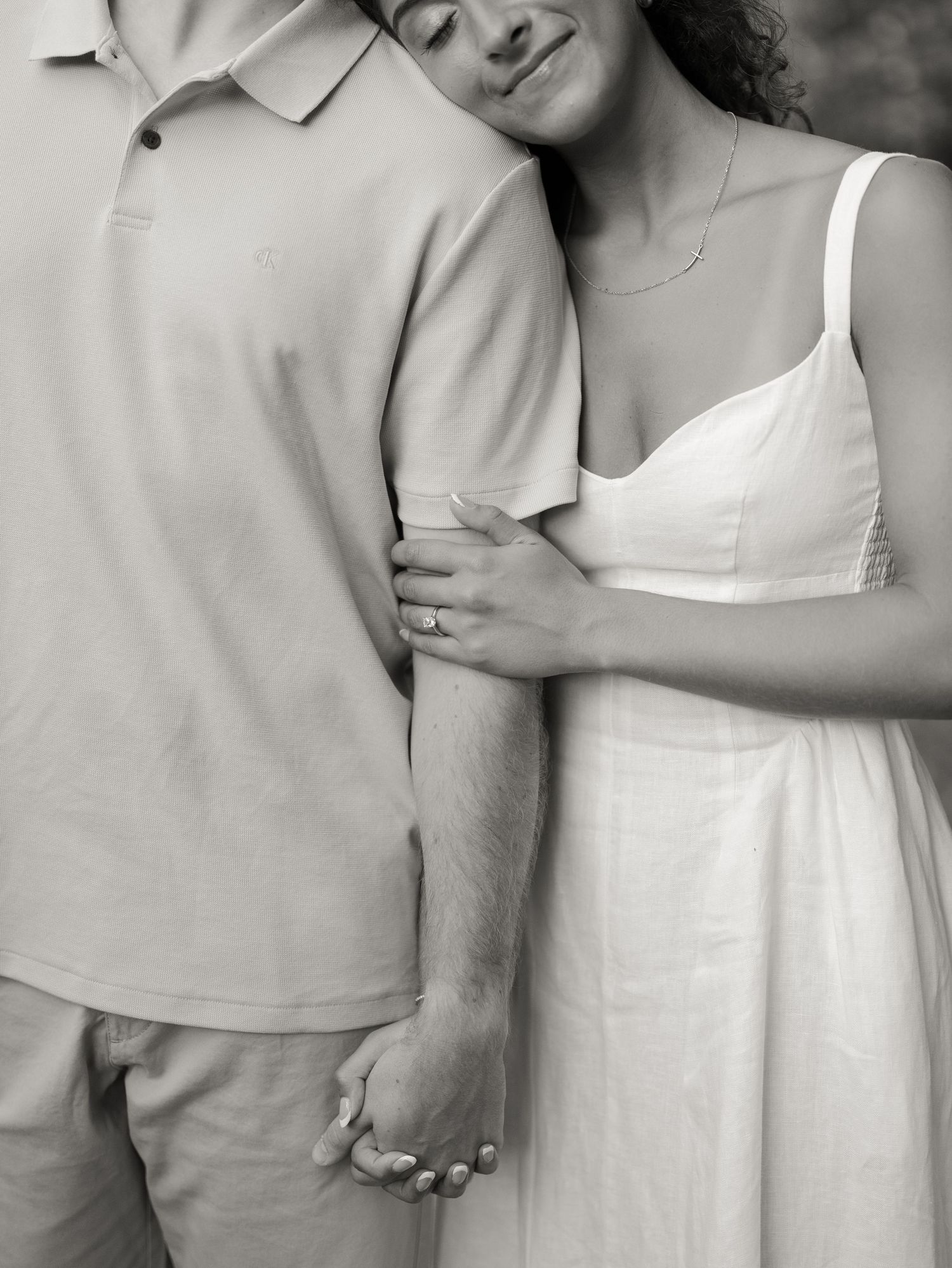 Black and white photo of a couple holding hands, woman in white dress with engagement ring resting hand on mans arm.