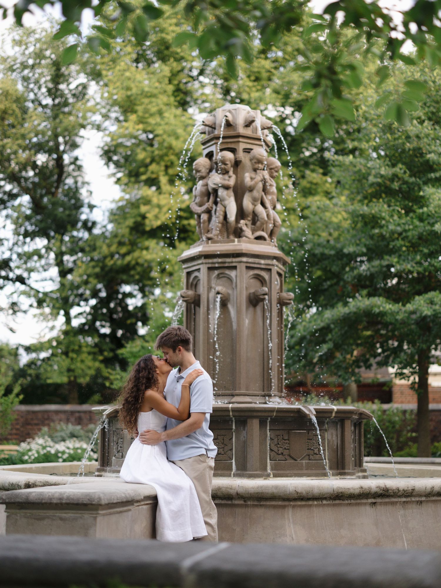 Couple sharing a romantic moment seated on an ornate stone fountain surrounded by lush greenery.