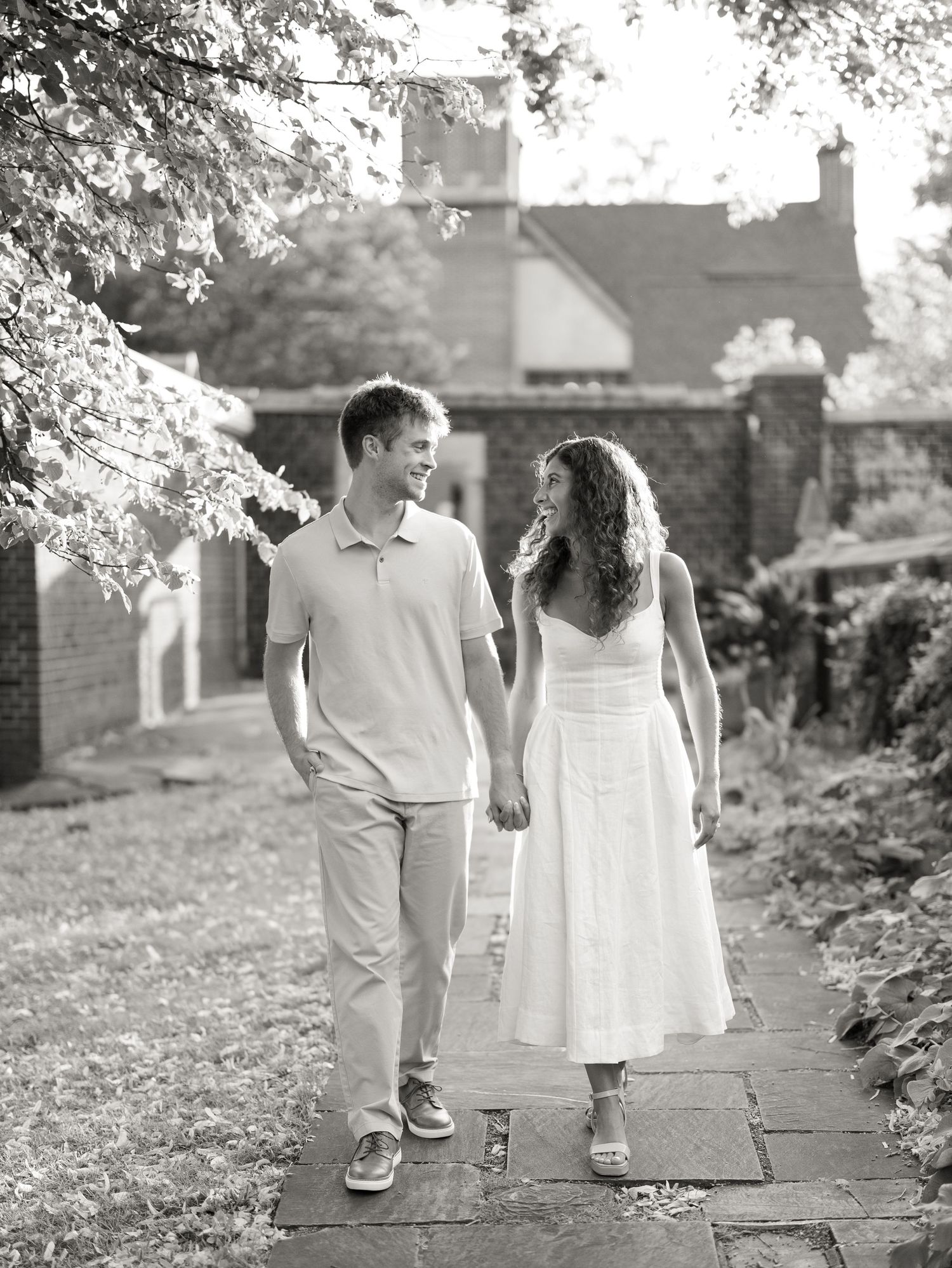 A couple holds hands and smiles while walking along a stone path surrounded by lush greenery in a black and white photo.