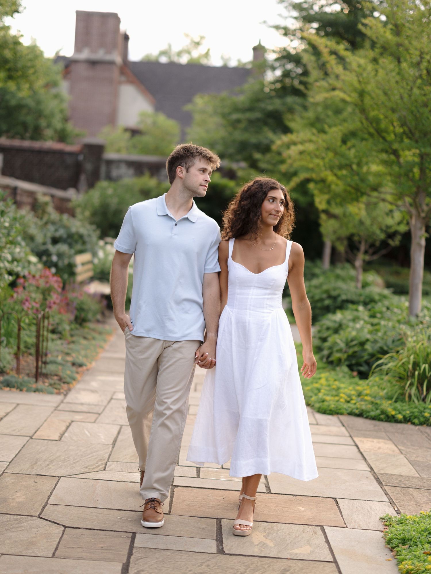 Couple holding hands walking on stone path in lush garden, man in blue polo and woman in white dress.