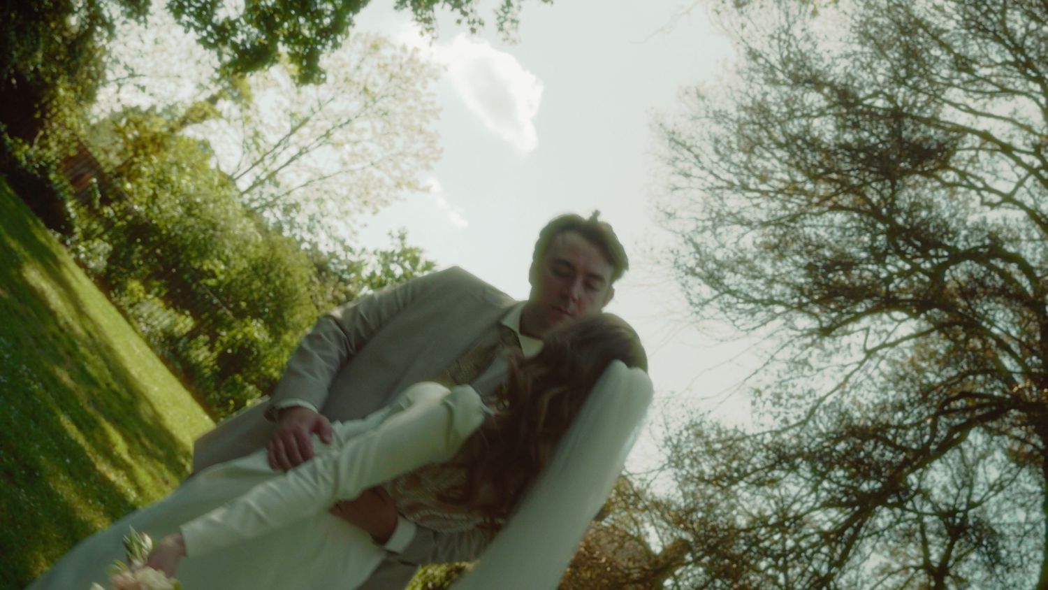 Man in suit carries woman in white dress outdoors in sunny green garden, low-angle artistic wedding photo.
