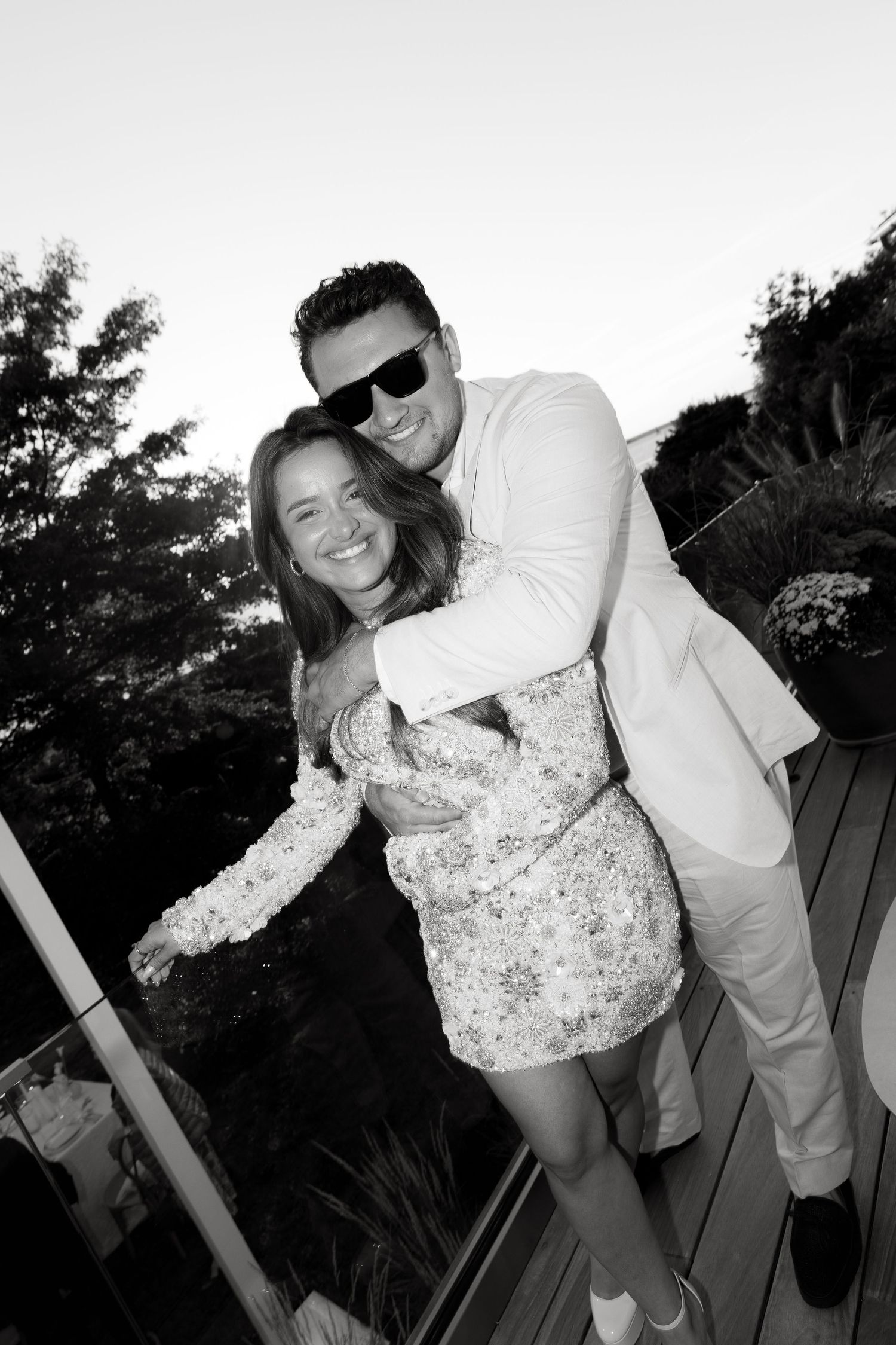 Black and white photo of a smiling couple embracing on an outdoor deck surrounded by trees at dusk.